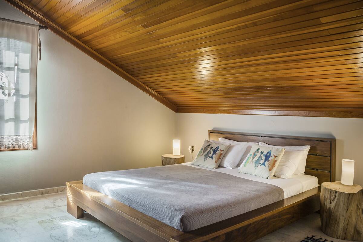 A bedroom with a wooden ceiling, a bed with a grey bedspread, and two bedside tables with lamps.