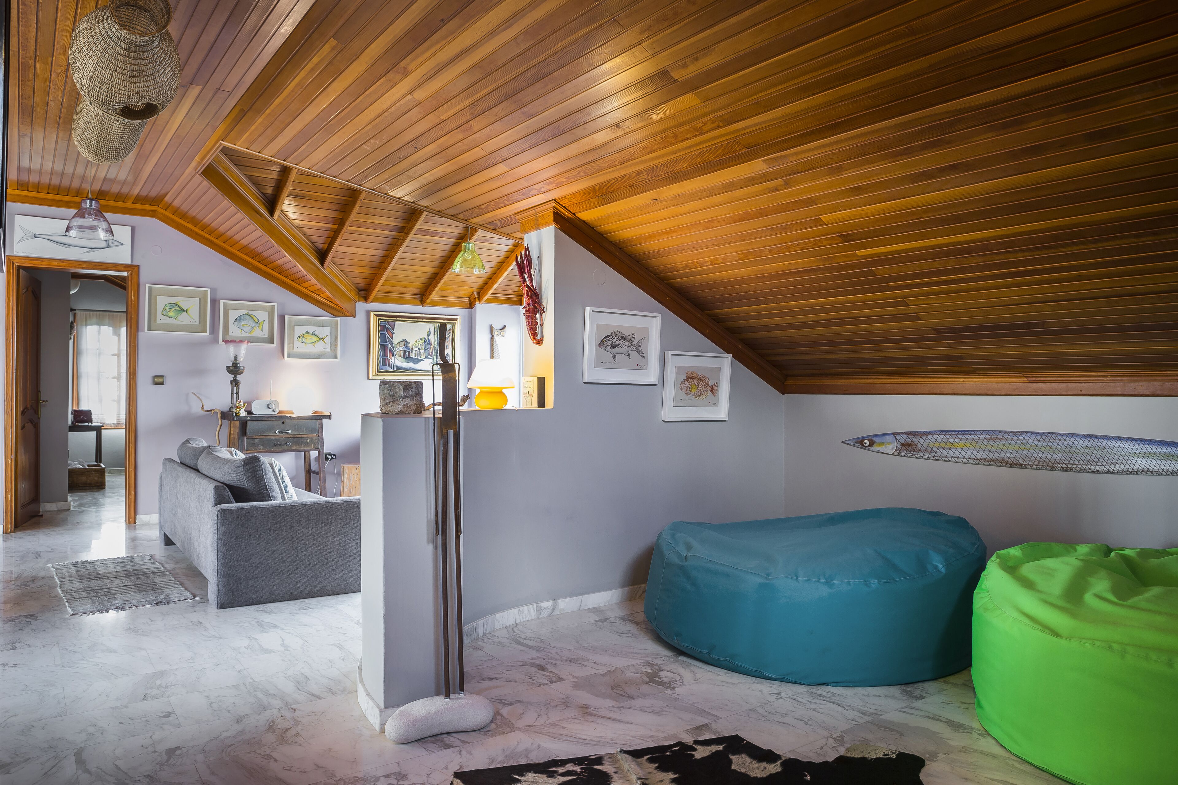 A room with a sloped ceiling, wooden beams, a sofa, a bean bag, a lamp, and framed pictures on the walls.