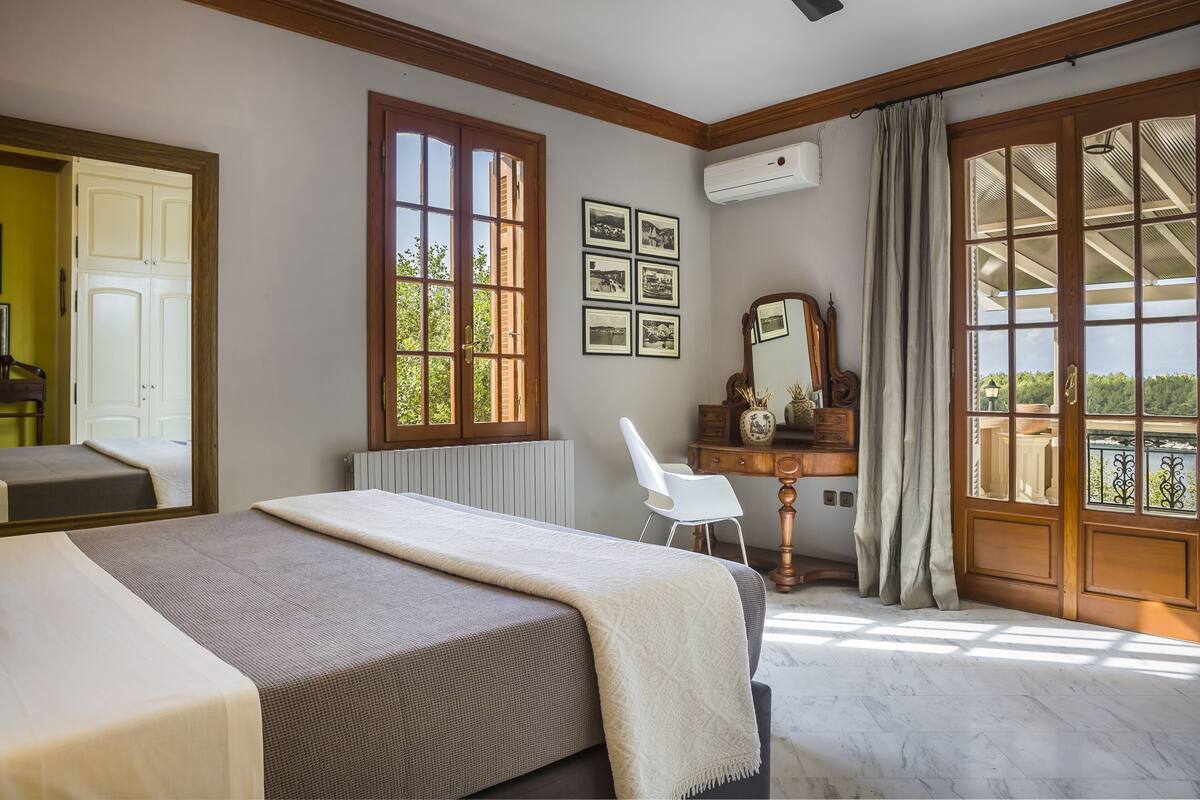 A bedroom with a bed, a desk, a chair, a mirror, and a window with a view.