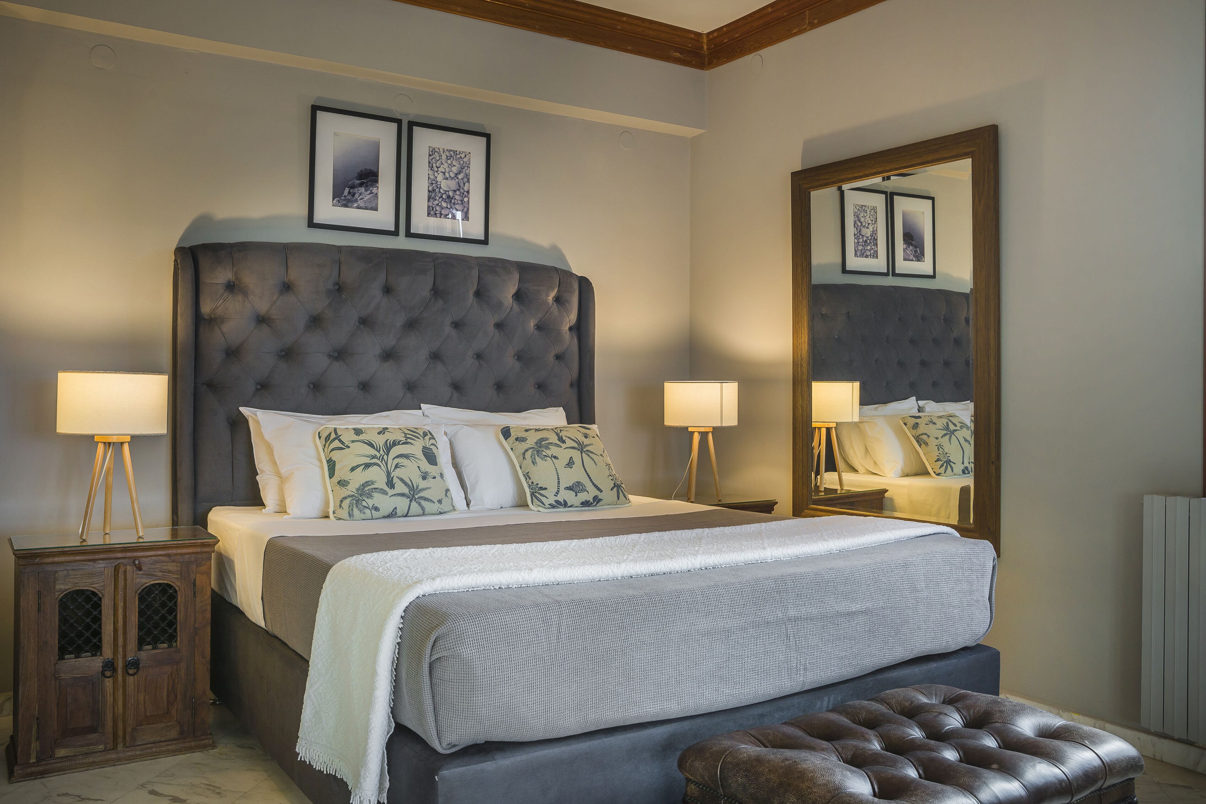 A bedroom with a large tufted headboard, two bedside lamps, a wooden nightstand, a large mirror, and a bench.