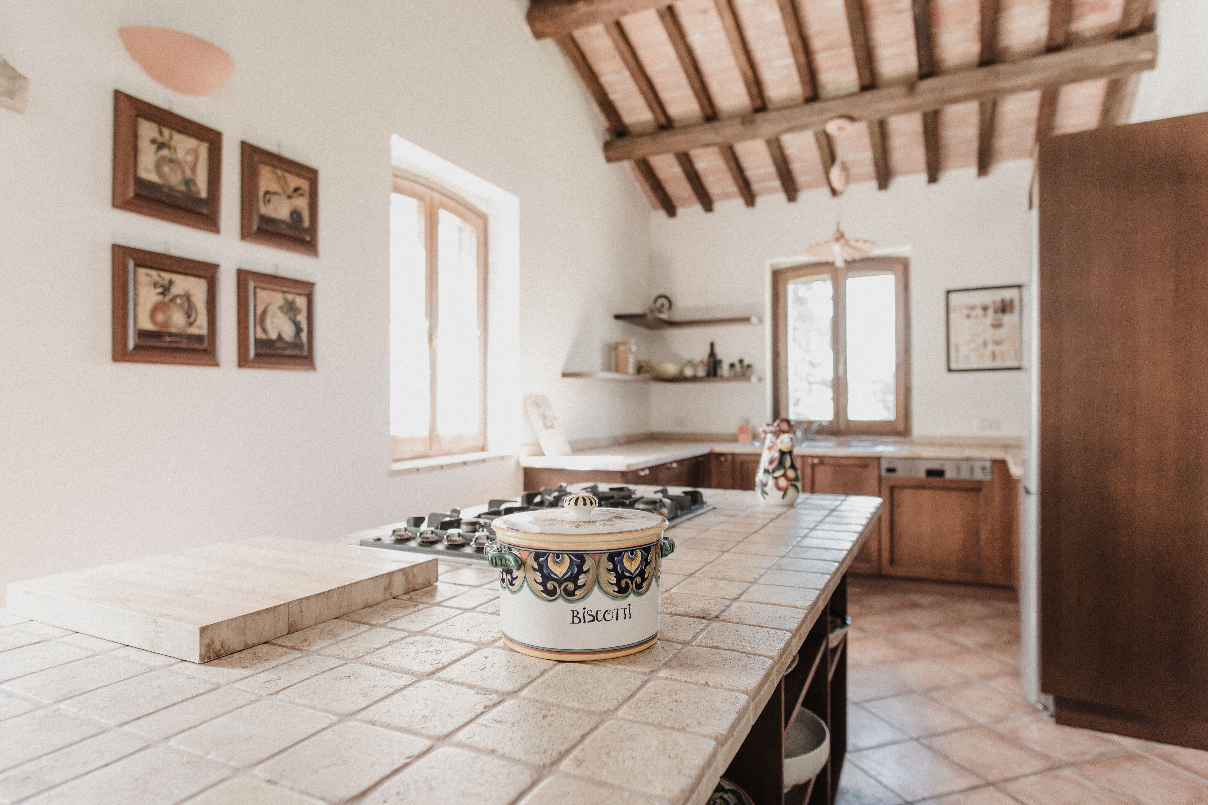 Una cocina con encimera de azulejos, estufa y una olla grande con el rótulo "Biscotti". Se observan cuadros en la pared y una ventana que deja entrar luz natural.