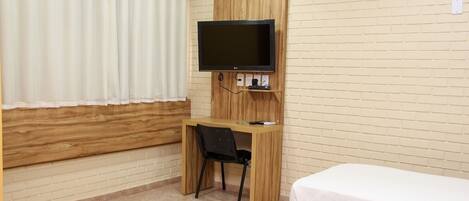 Habitación de hotel con un escritorio de madera, un televisor montado en la pared, una cama con colcha blanca y una ventana con cortinas blancas.