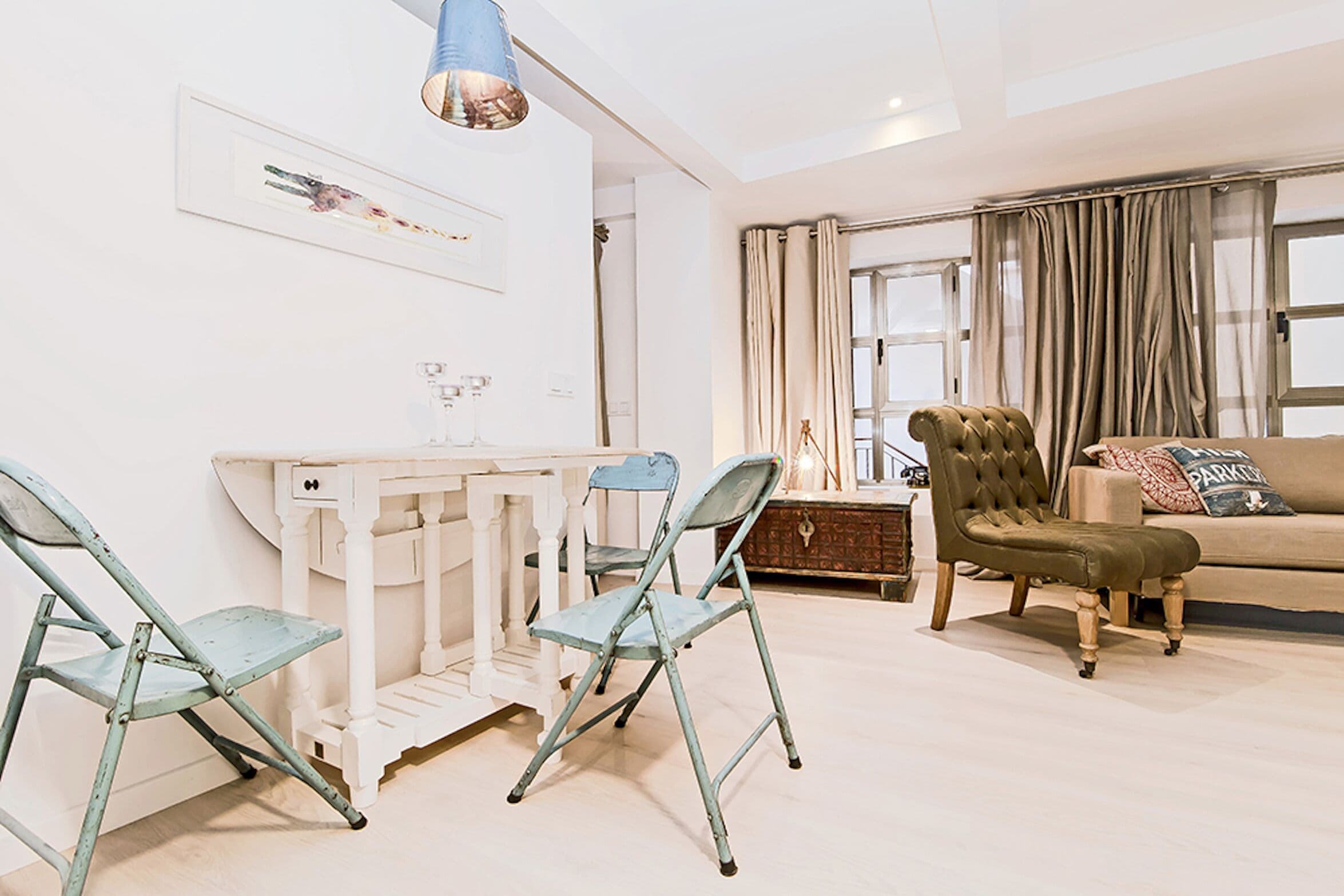 Een woonkamer met een witte bank, een groene fauteuil en een blauwe opklapbare stoel. Er staat een witte tafel met een glazen blad en er hangt een blauwe lamp.