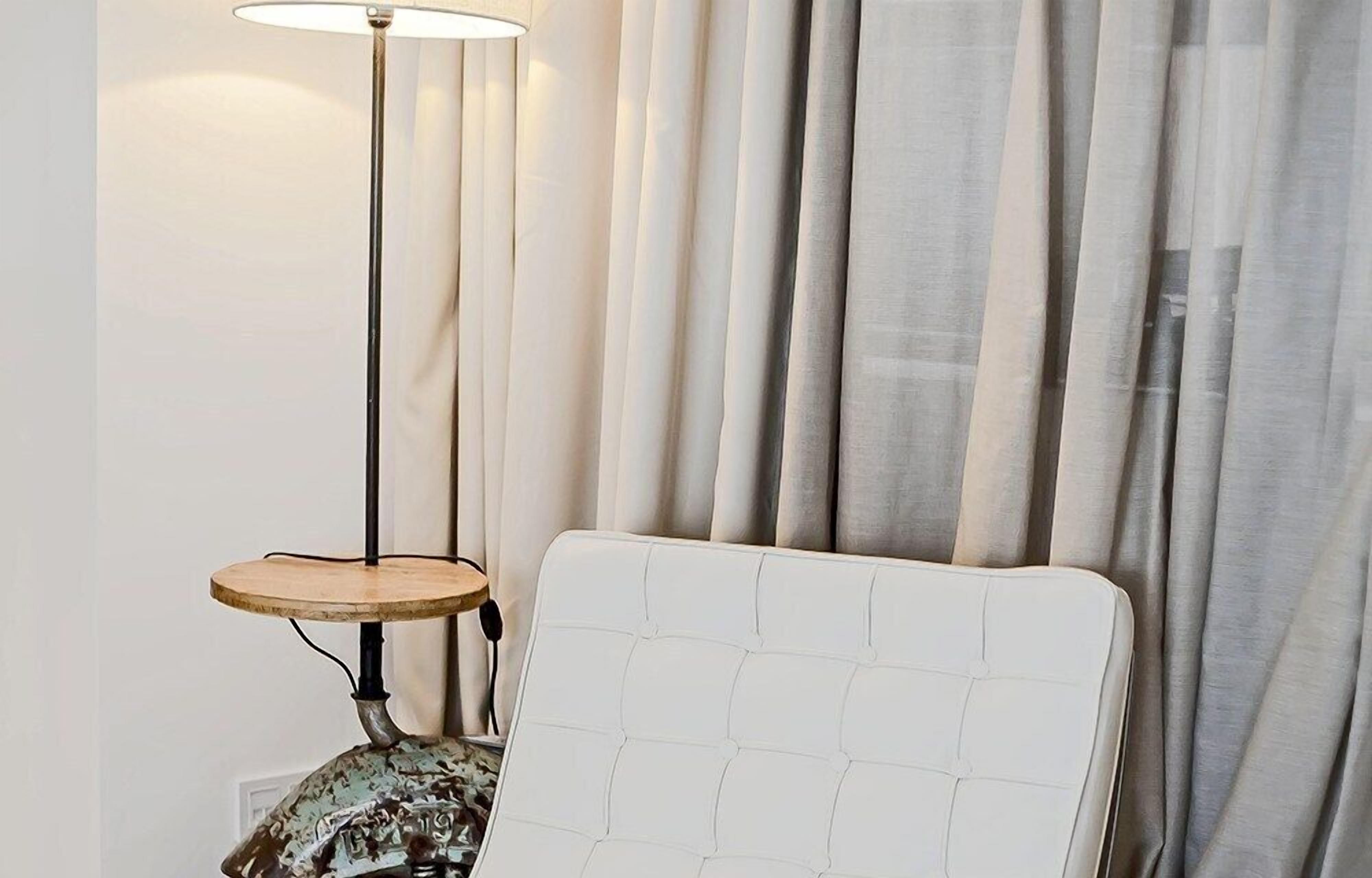 Une pièce avec un fauteuil recouvert de tissu blanc, une petite table ronde avec une lampe et un rideau en arrière-plan.