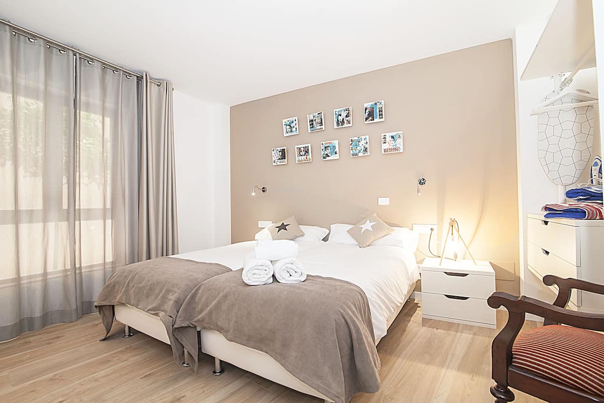 Un dormitorio ordenado con cama, mesitas de noche, una silla y una pared decorada con cuadros.