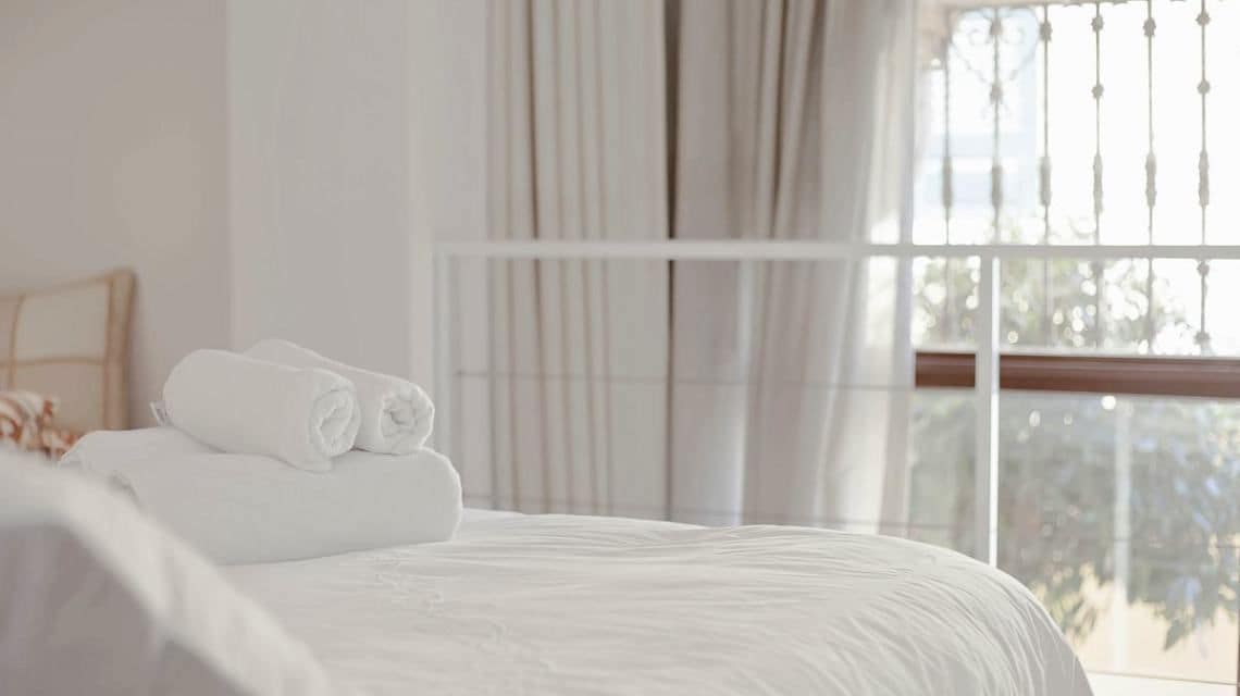 A neatly made bed with rolled white towels, a window with vertical blinds, and a view of trees outside.