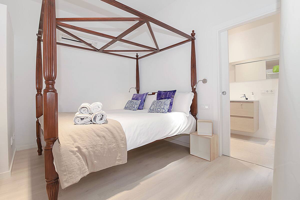 Una cama con dosel de madera, cubrecama blanco y almohadas azules con estampado. Hay toallas enrolladas sobre la cama.