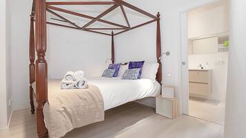 Ein Bett mit Baldachin, dessen Gestell aus Holz gefertigt ist, mit weißer Bettdecke und blumengemusterten Kissen. Auf dem Bett liegen gerollte Handtücher.
