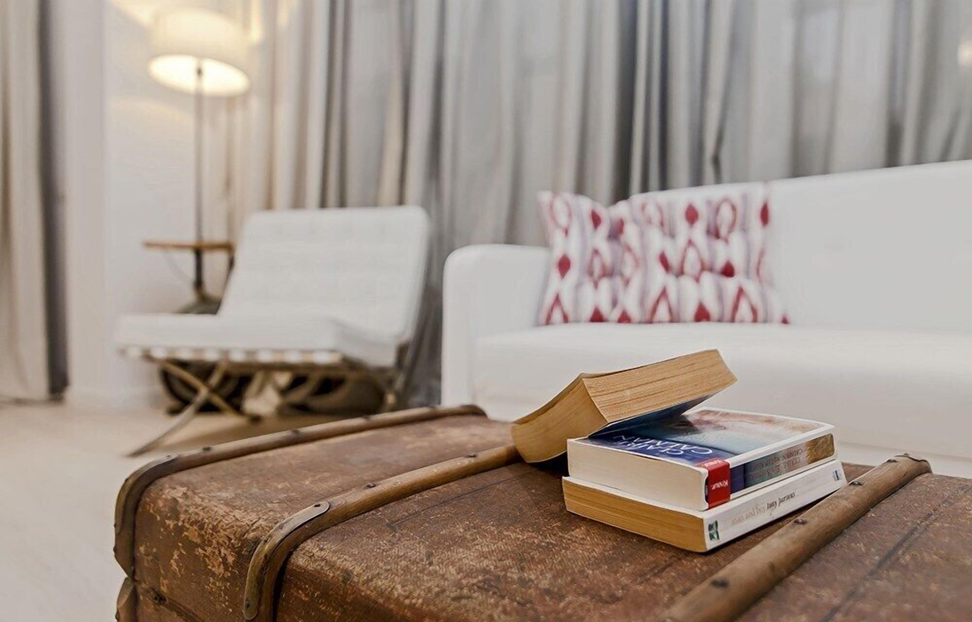 Een woonkamer met een witte bank, een gekleurd kussen, een houten tafel met boeken en een vintage koffer.
