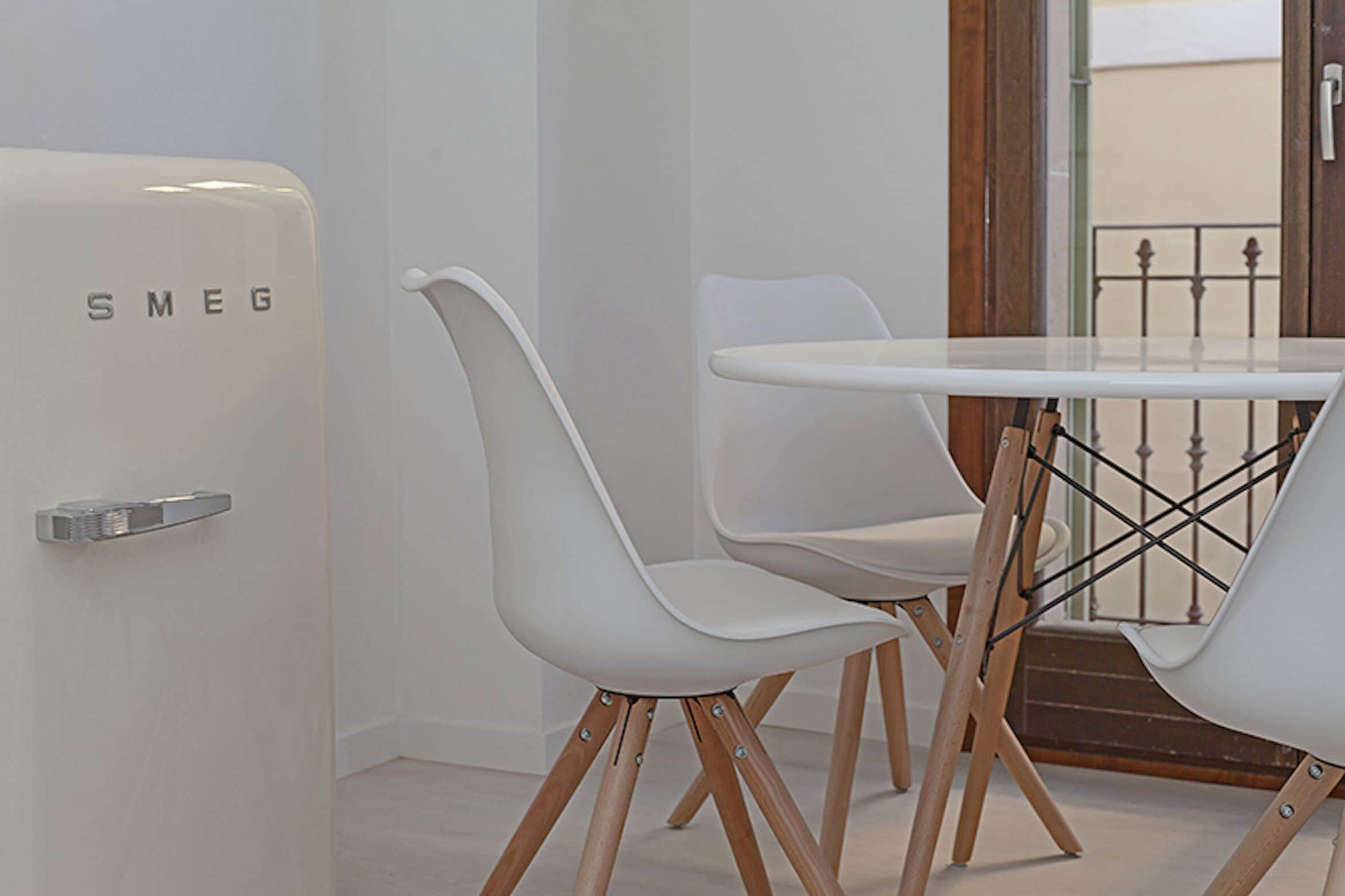 Une pièce moderne avec une table et des chaises blanches, un mur blanc et une porte portant l’inscription « SMEG ».
