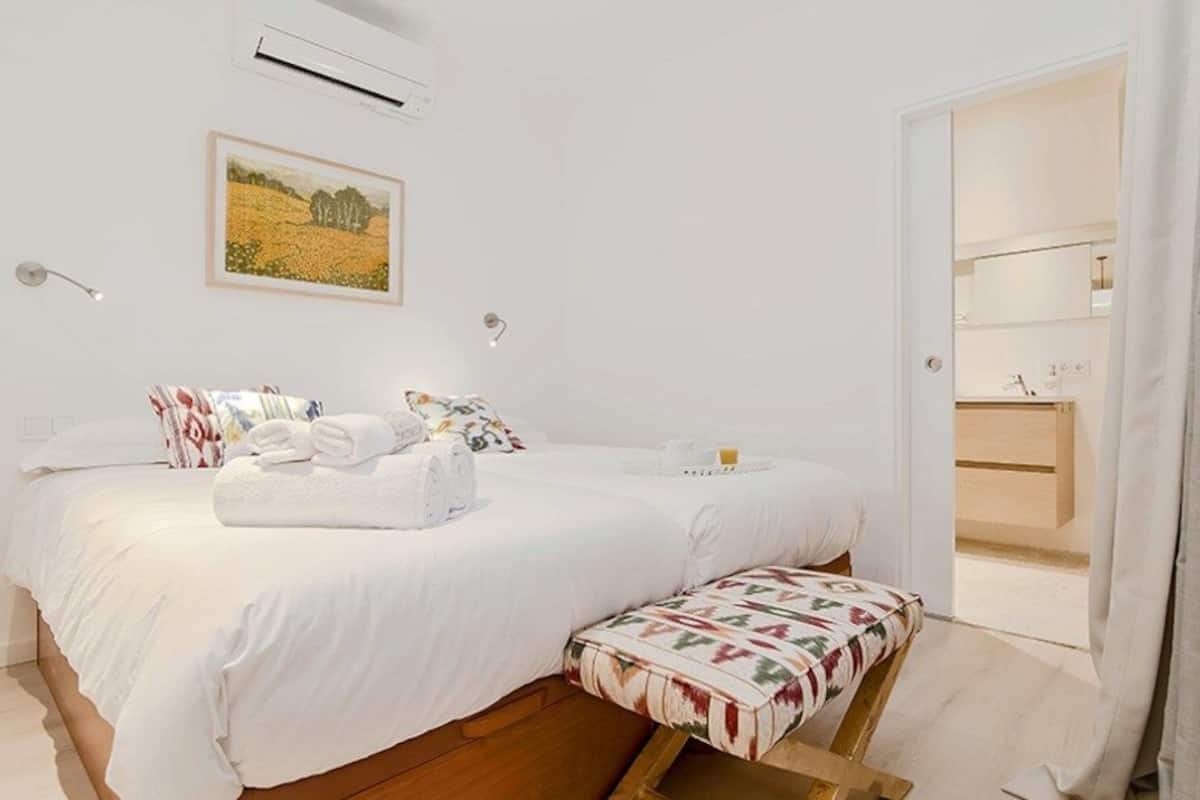 Un dormitorio con cama, una banca, un cuadro y un baño visible a través de una puerta abierta.