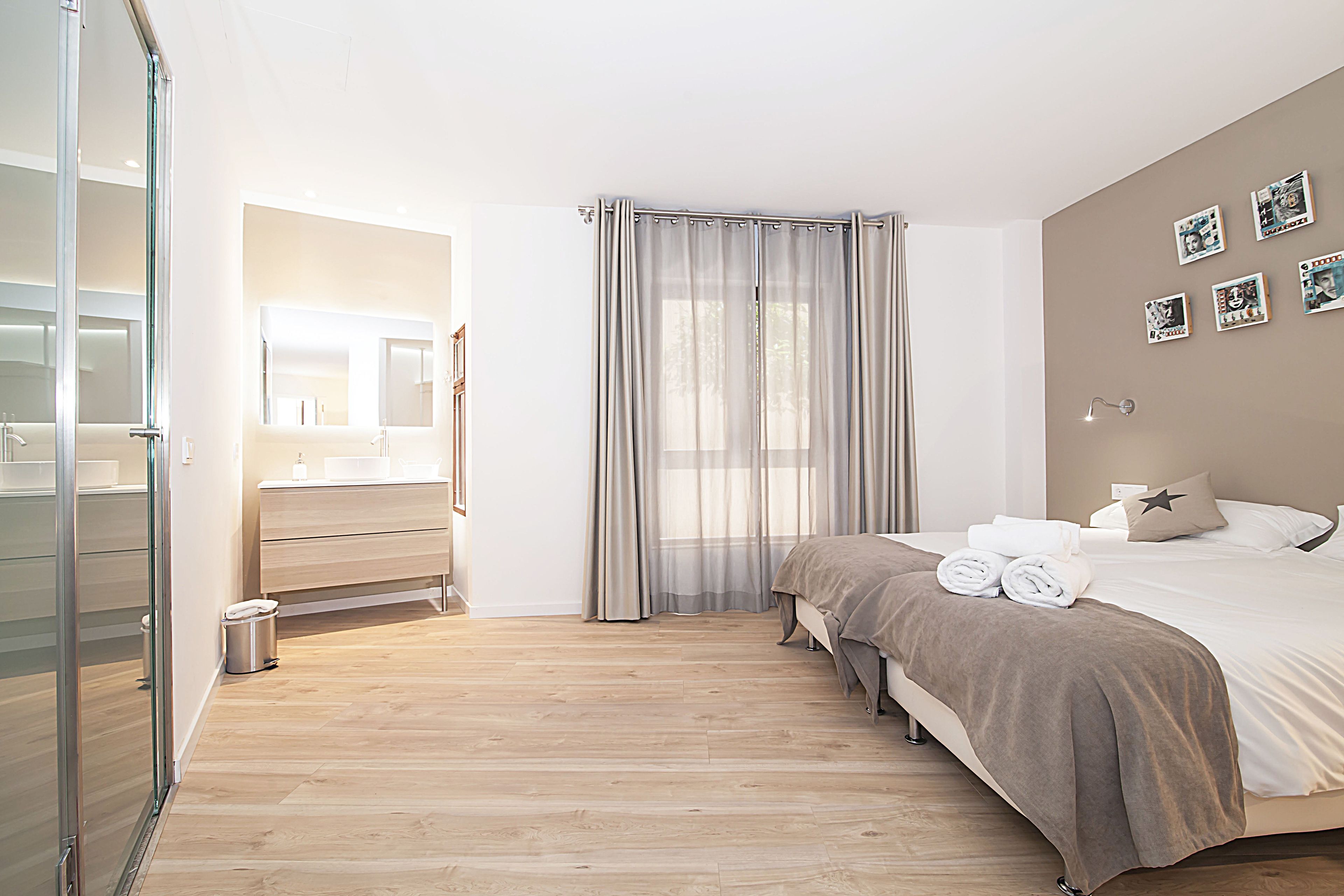 Een moderne hotelkamer met een houten vloer, een bed met een beige deken, een nachtkastje, een spiegel en een raam met dunne gordijnen.