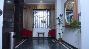 A hotel lobby with a marble floor, two red chairs, a small table, a potted plant, a mirror, and a window with curtains. - Sea Shell Hotel (Dhaka)