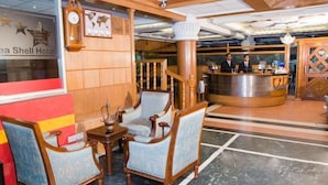 Um saguão de hotel com uma mesa de recepção, dois recepcionistas, um relógio e uma área de estar com cadeiras e uma mesa.