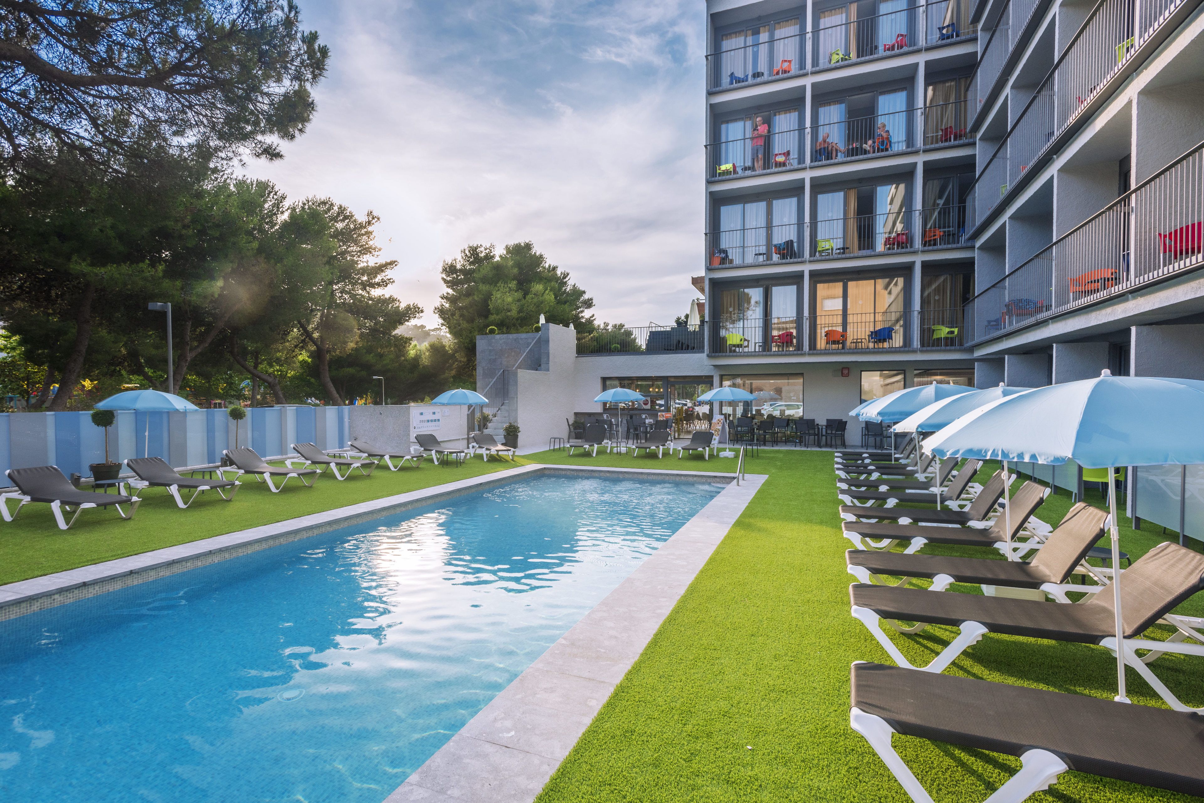A swimming pool with sun loungers and umbrellas, surrounded by a building with balconies and a green lawn.