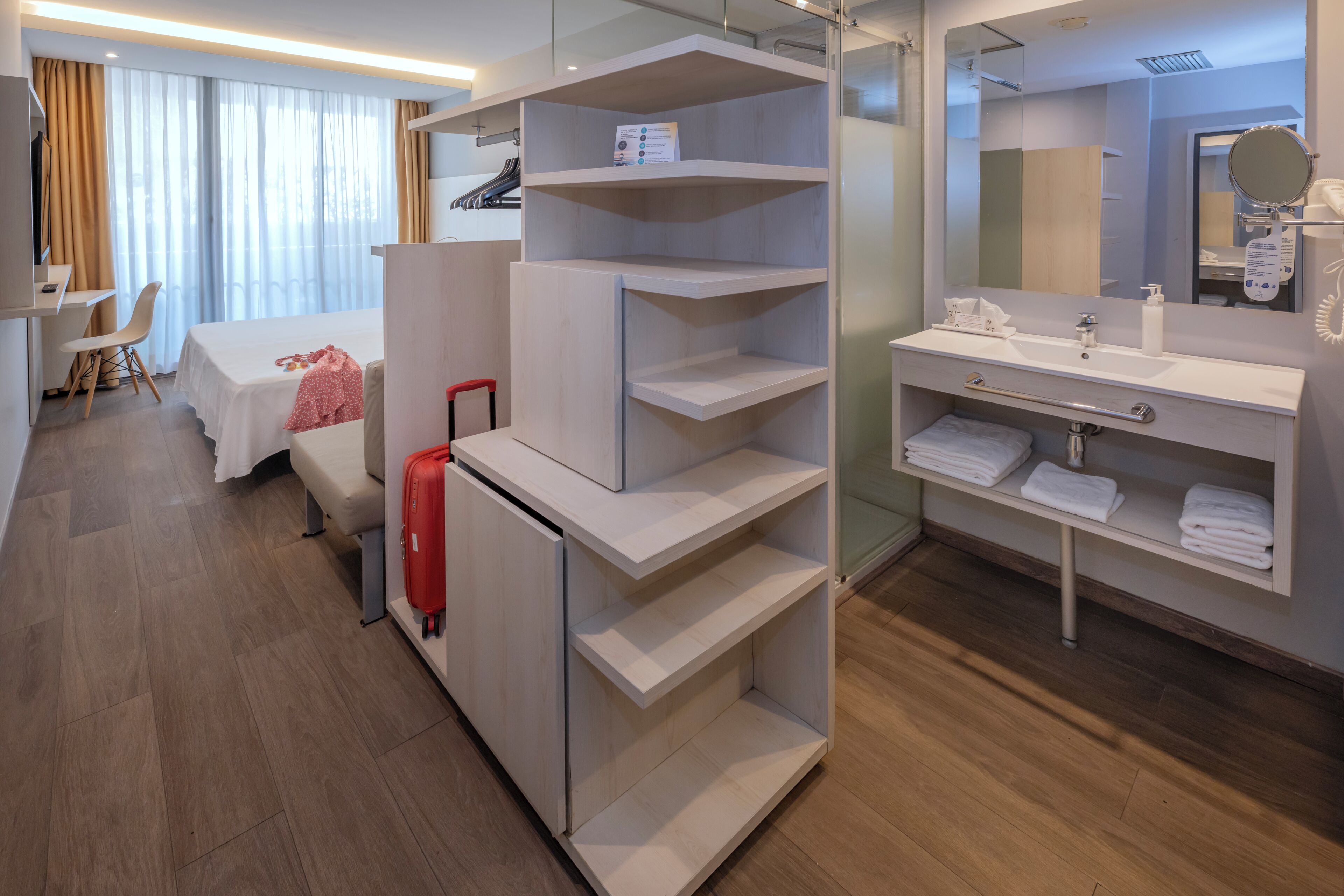 Una habitación de hotel con una cama, un escritorio, una silla, una maleta roja, un tocador con espejo y estantes con toallas.