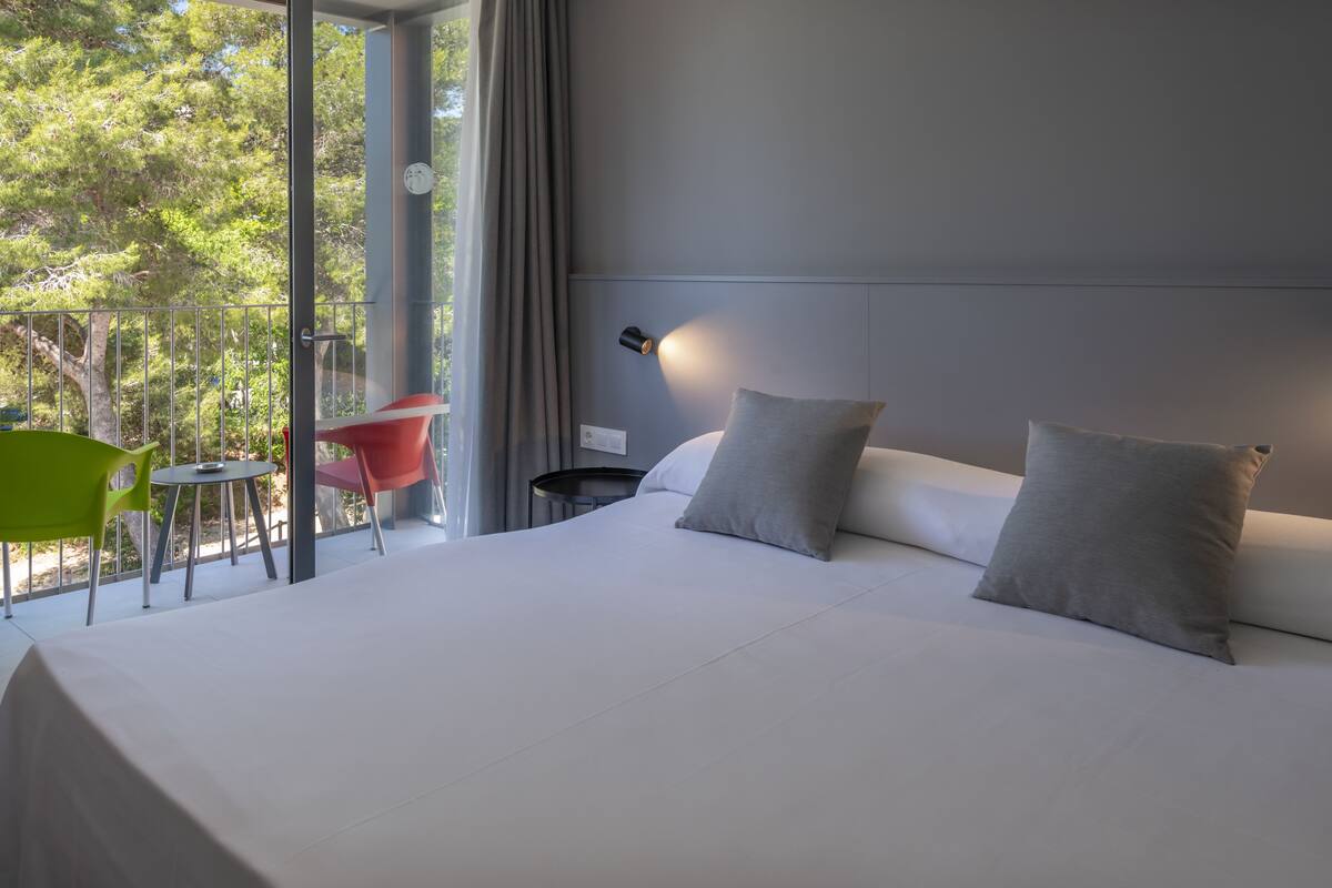 Una habitación de hotel moderna con una cama grande, dos almohadas, una mesita de noche, una lámpara y vistas a árboles a través de amplias ventanas.