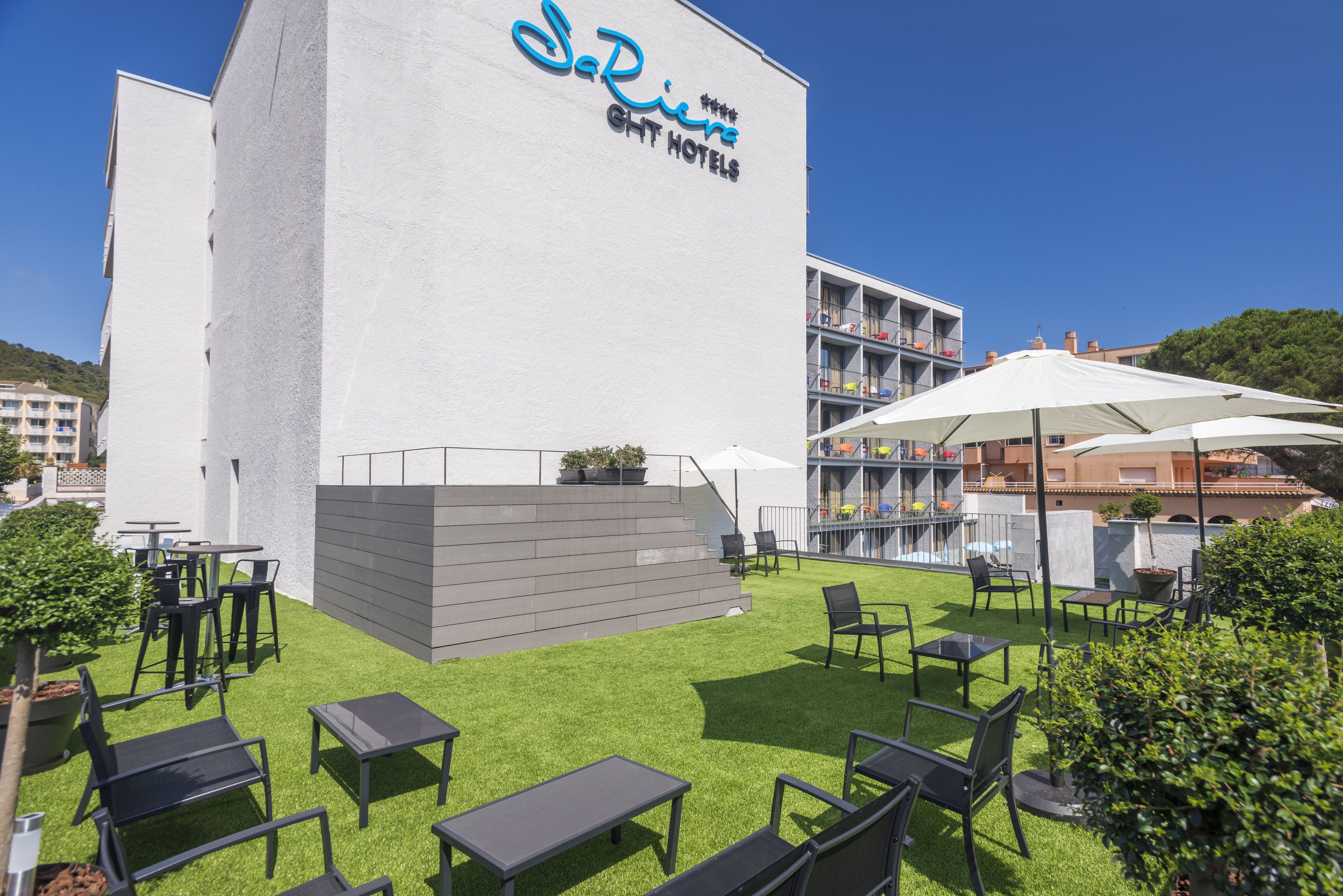 Ein Hotel mit weißer Fassade, Außenbereich mit Tischen und Stühlen, und einem strahlend blauen Himmel.