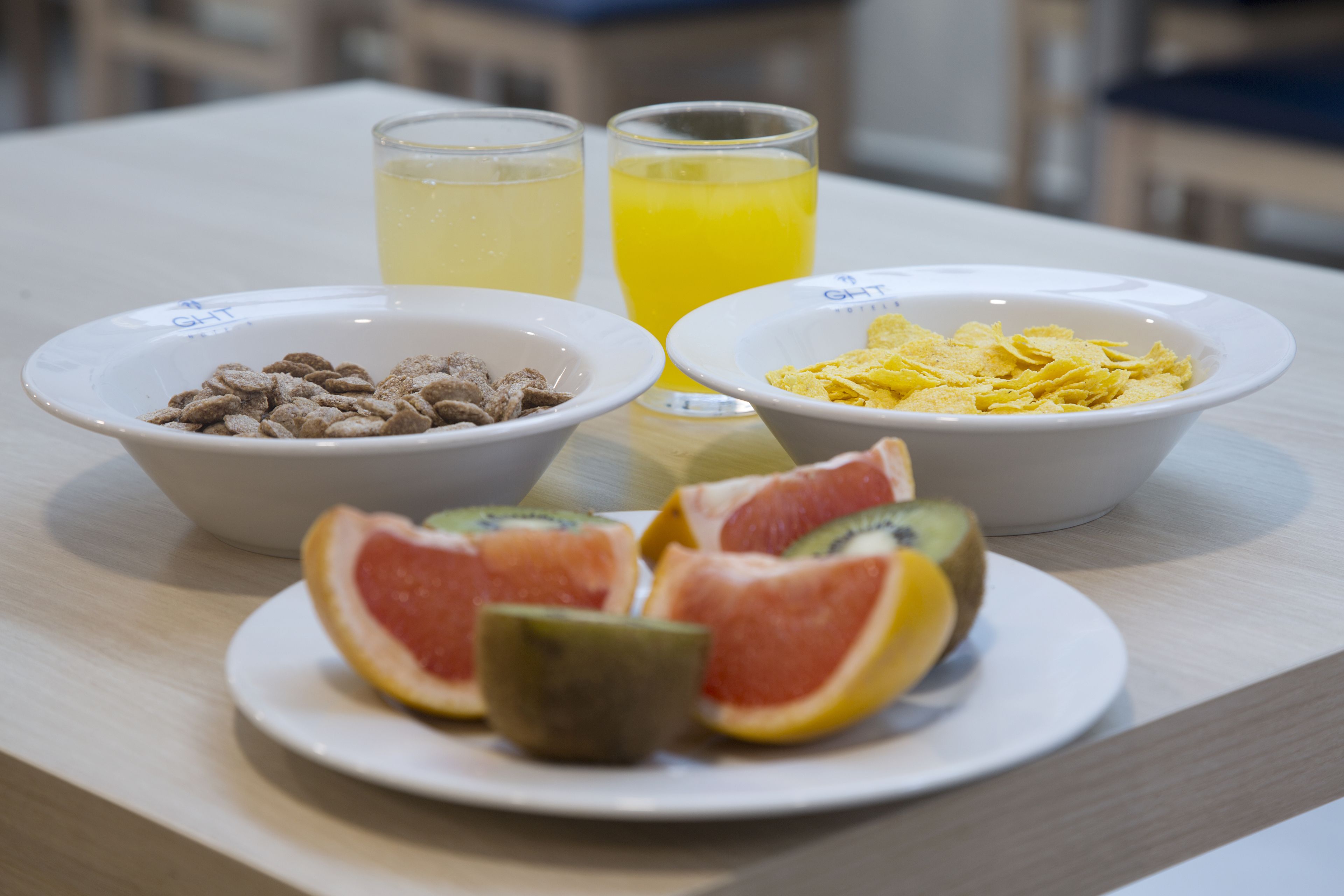 A breakfast table with bowls of cereal, a plate of sliced fruit, and two glasses of orange juice.