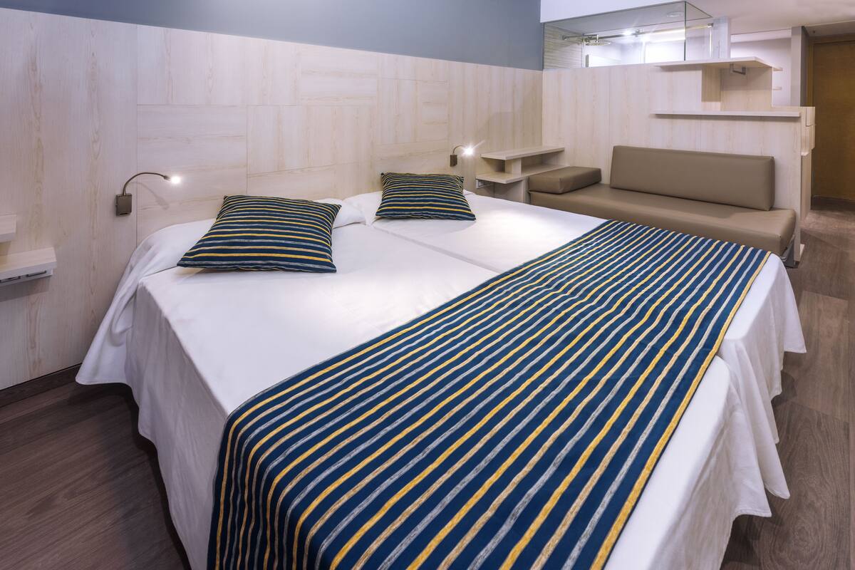 Una habitación de hotel con una cama grande, dos almohadas, una mesita de noche y un sofá, junto con una pequeña mesa con lámpara.