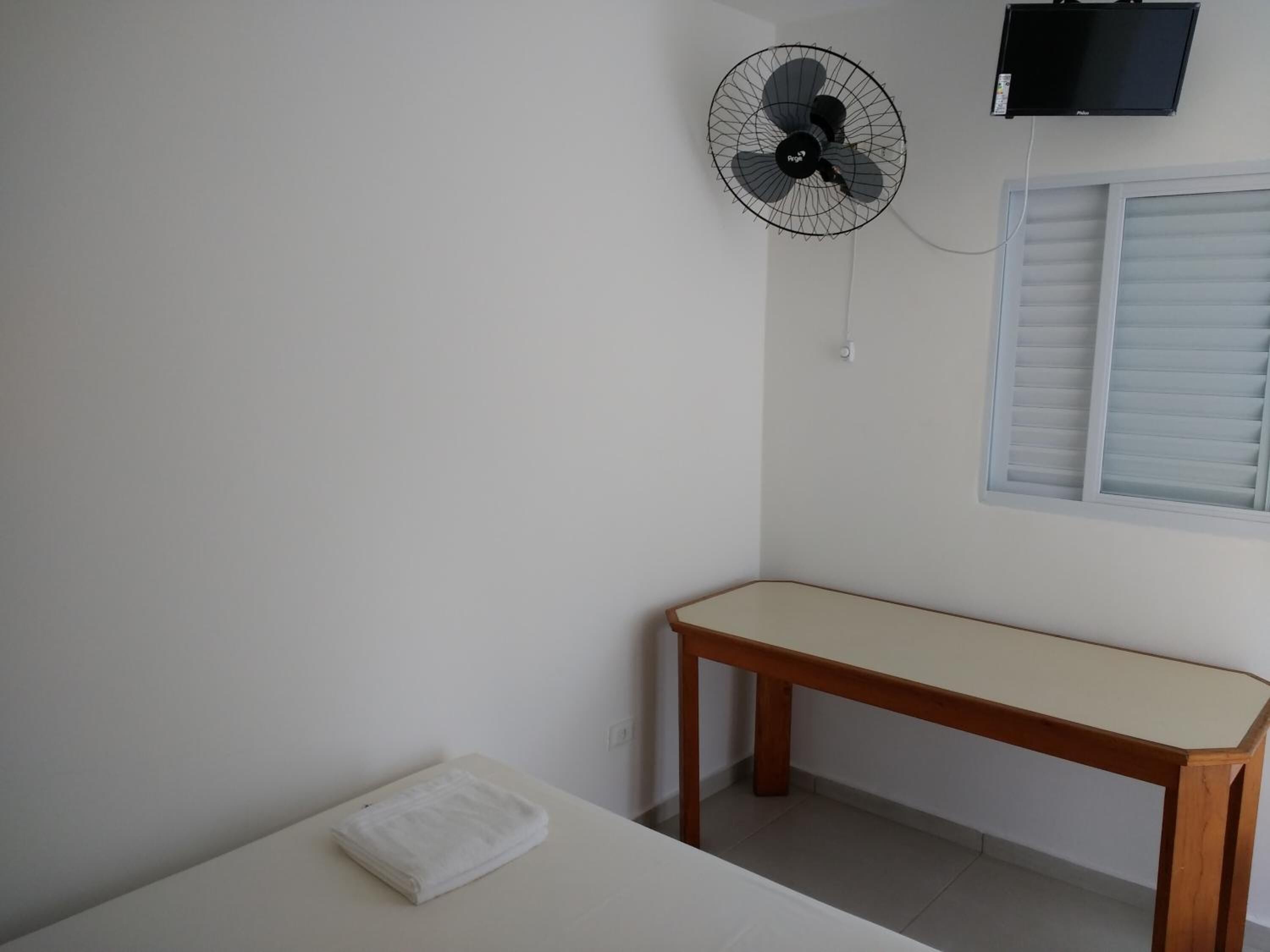 A room with a bed, a wooden table, a wall-mounted fan, and a TV.
