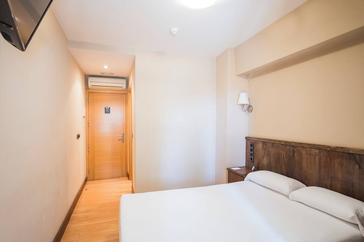 Een hotelkamer met een houten vloer, een bed met witte lakens, een nachtkastje, een wandlamp en een deur die naar een andere kamer leidt.