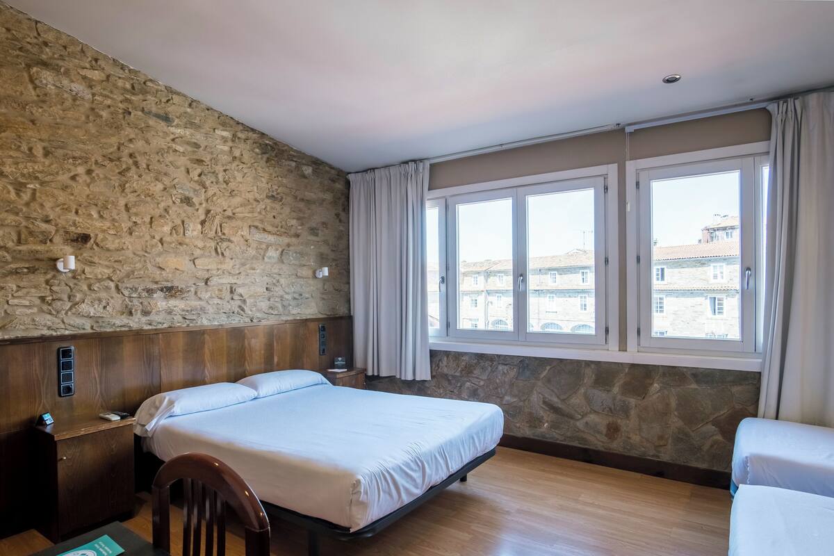 Een hotelkamer met een stenen muur, een houten hoofdbord, een bed met witte lakens, een houten nachtkastje, een bureau met stoel, en een raam met gordijnen.
