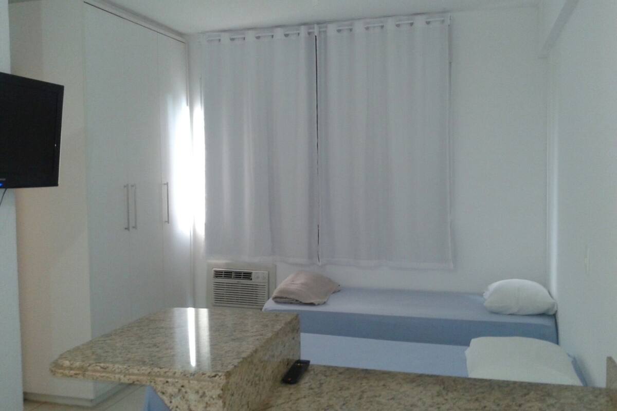 Une petite chambre propre, avec un lit, un téléviseur fixé au mur et une fenêtre avec des rideaux.