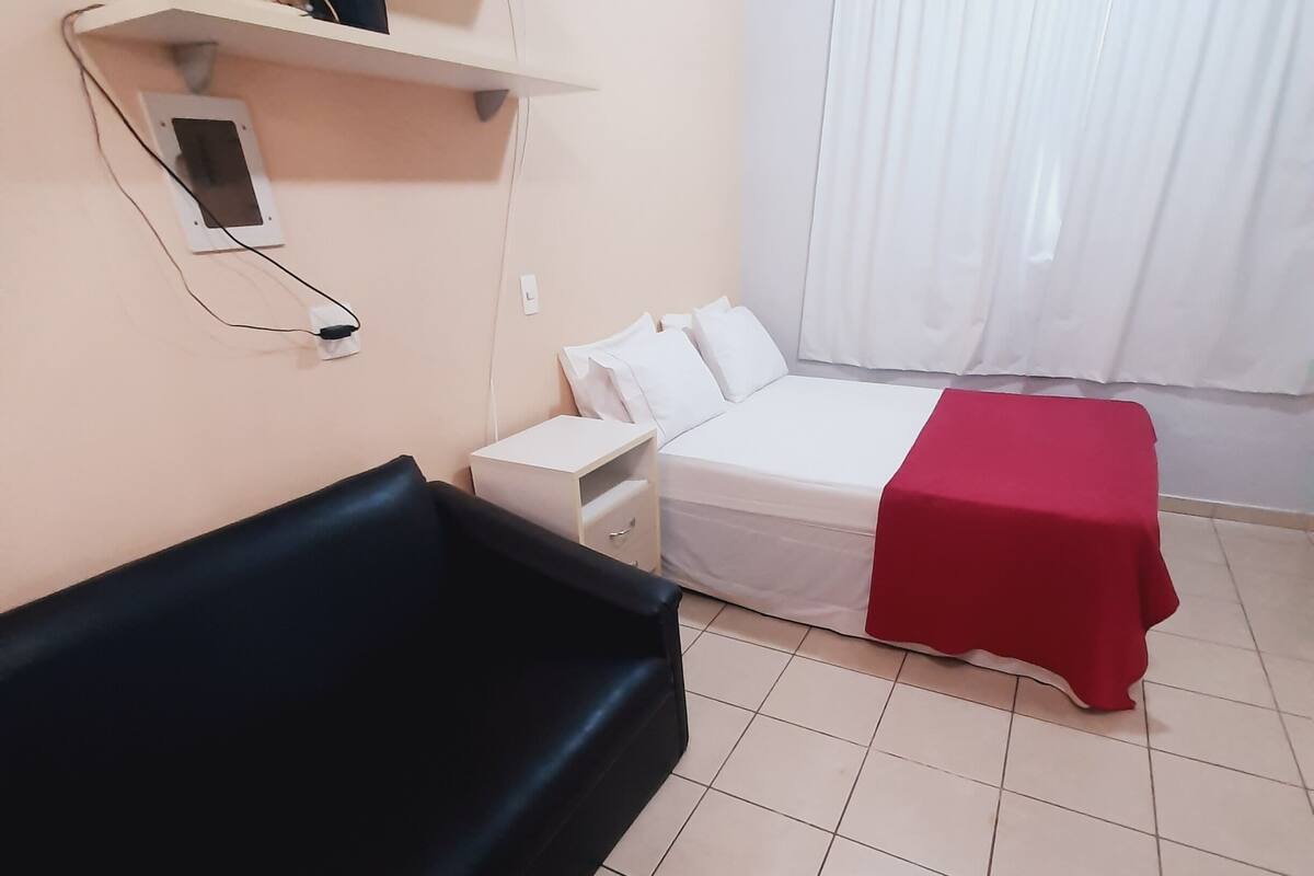 Une chambre d’hôtel avec un lit, une table recouverte d’une nappe rouge, un canapé noir, une table de chevet blanche, un climatiseur fixé au mur et une prise de courant.
