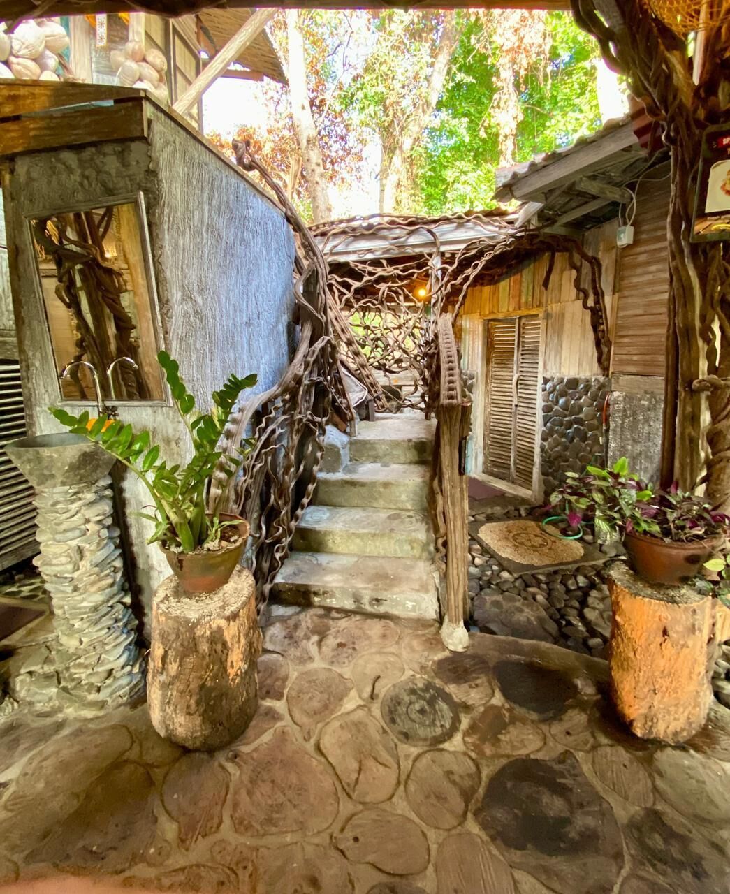 Una escalera rústica con escalones de piedra y barandillas de madera, rodeada de plantas en macetas y una estructura de madera con techo de paja.