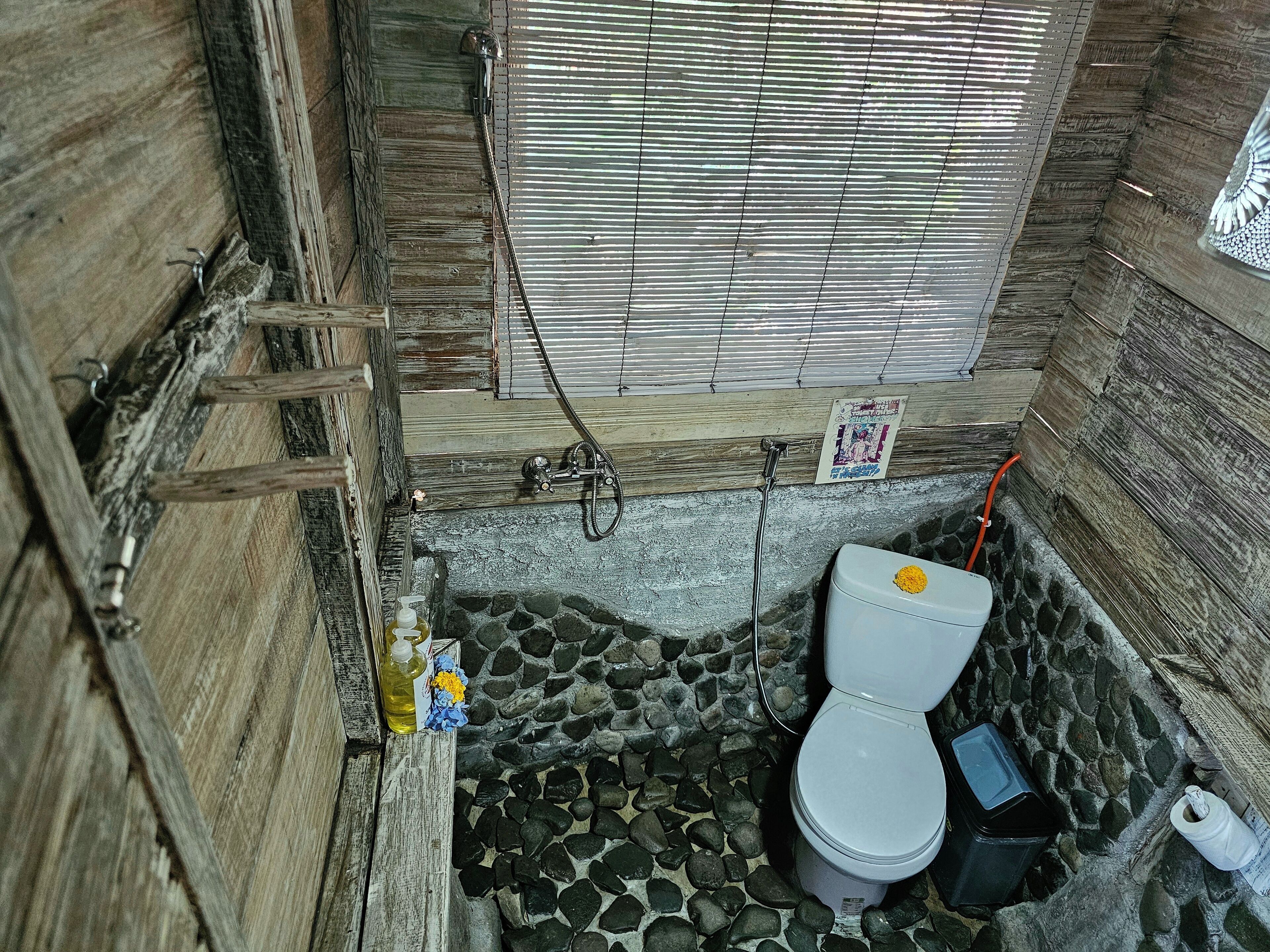 A rustic bathroom with a stone floor, wooden walls, a white toilet, and a shower area.