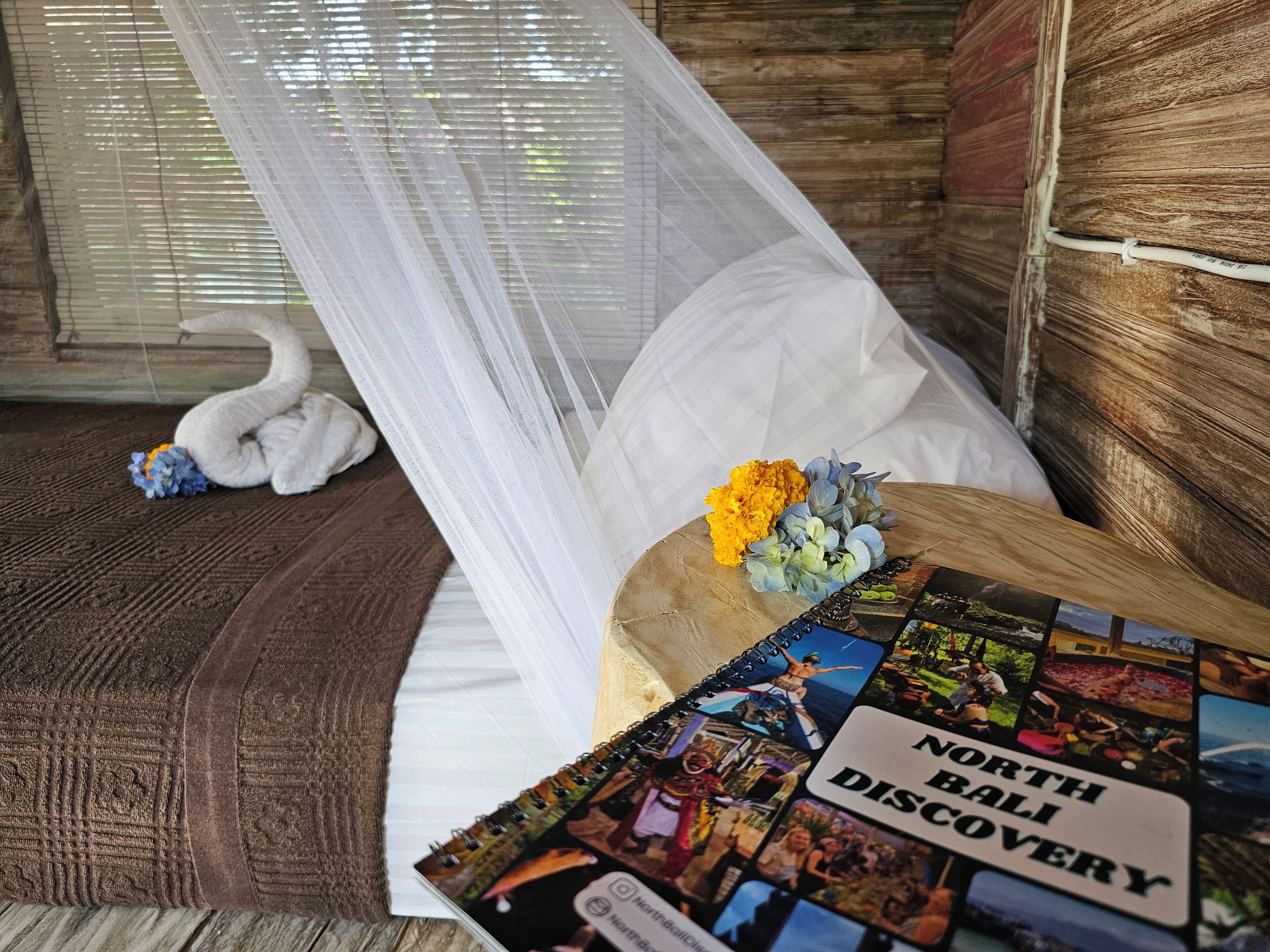 A room with a bed, a white swan decoration, a small bouquet of flowers, and a travel magazine titled "North Bali Discovery".