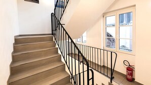 A staircase with a black railing, a fire extinguisher, and a window with a view of buildings. - Hotel Havana (Mainz)