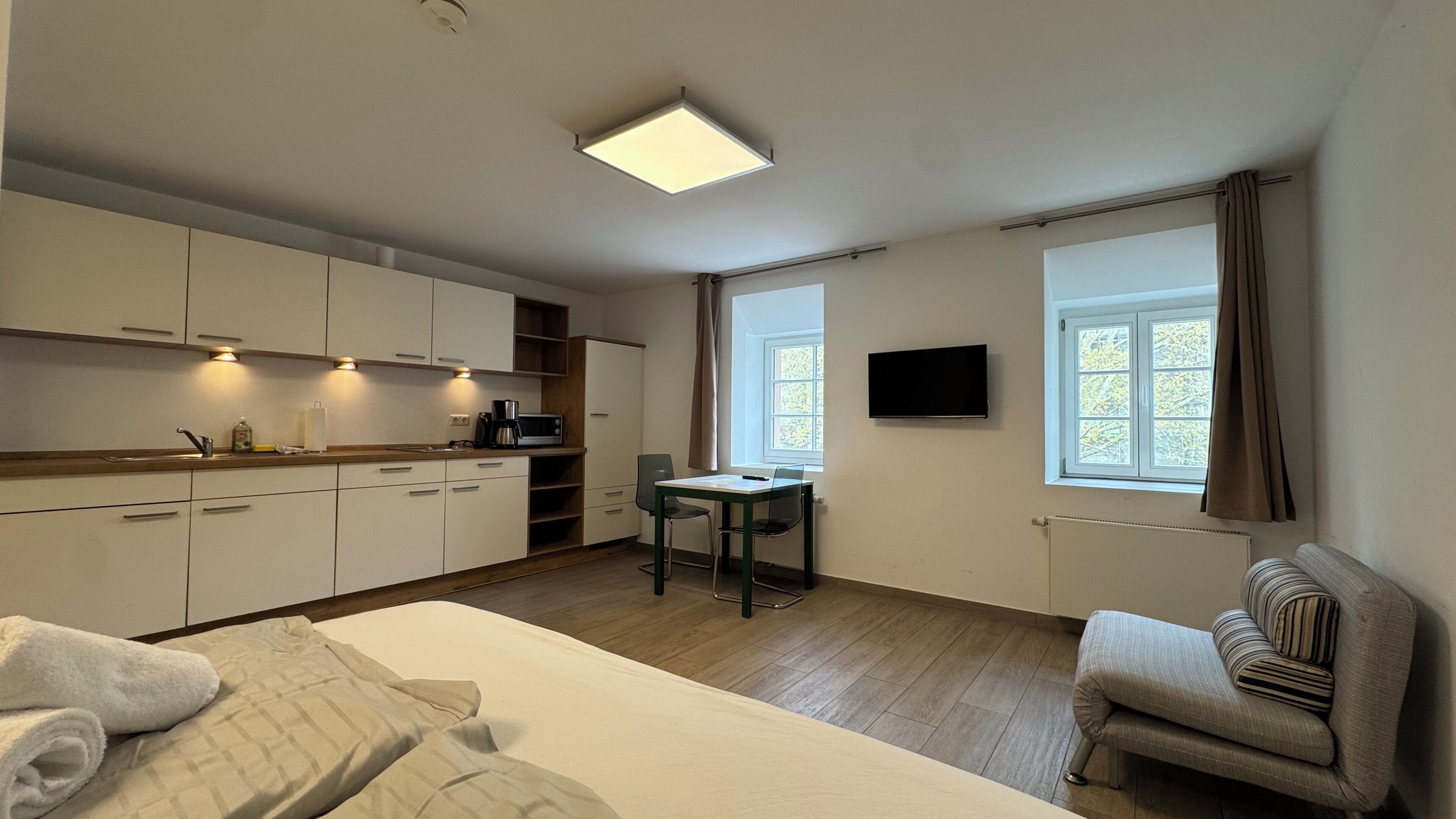Habitación de hotel compacta con kitchenette, una cama individual, una pequeña mesa, una silla, un televisor montado en la pared y una ventana con cortinas.