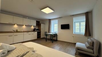 Habitación de hotel compacta con kitchenette, una cama individual, una pequeña mesa, una silla, un televisor montado en la pared y una ventana con cortinas.