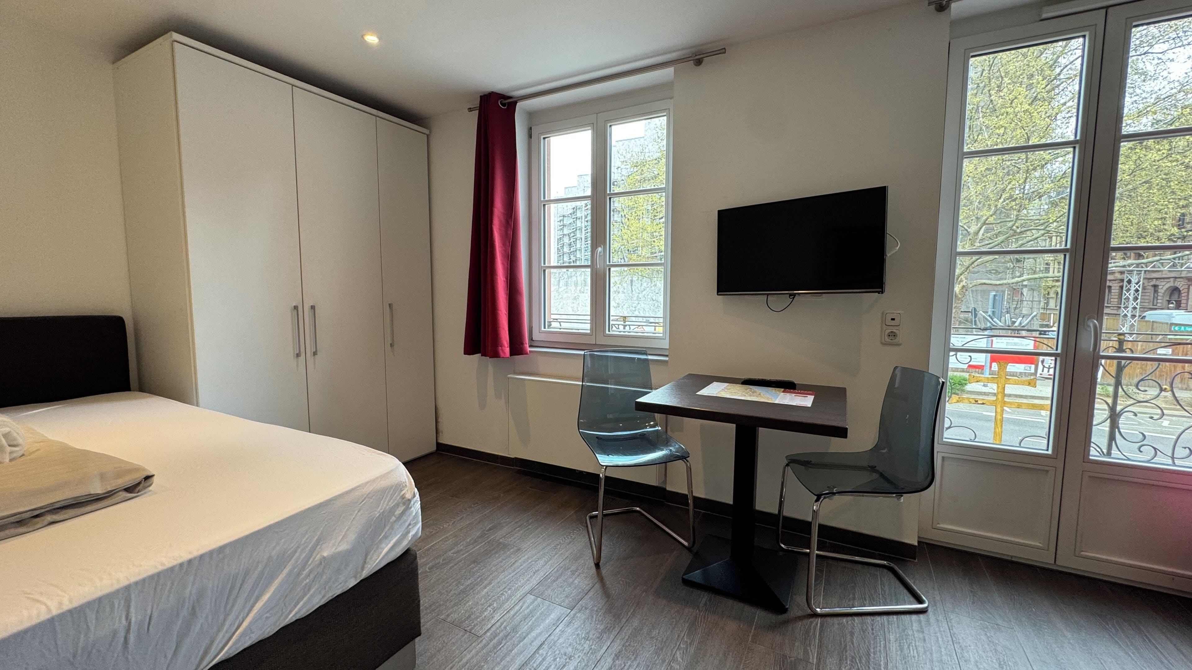 A hotel room with a bed, a desk with a chair, a TV, and large windows.