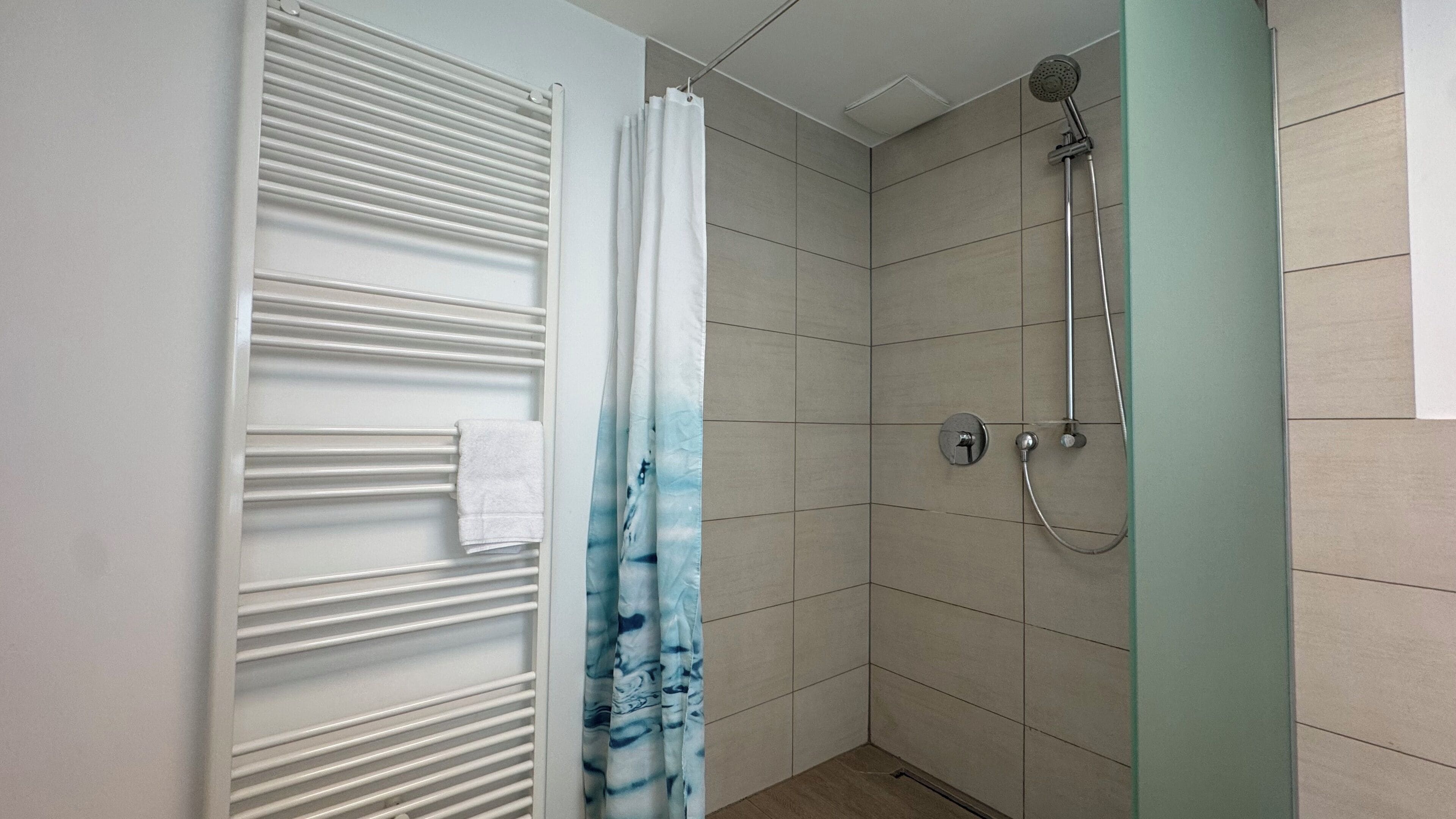 A shower with a handheld showerhead and a wall-mounted faucet, a towel rack with a towel, and a door with a white and blue curtain.