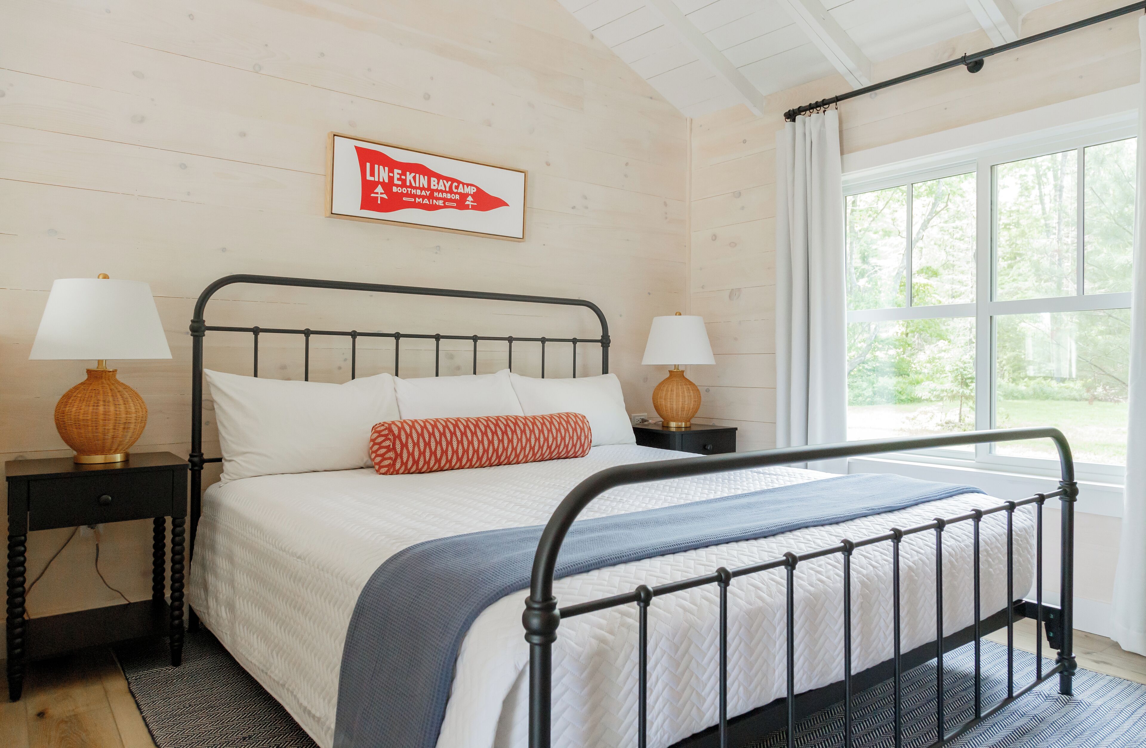 A bedroom with a metal bed frame, white bedding, two bedside tables with lamps, a framed sign on the wall, and a large window with a view of trees.