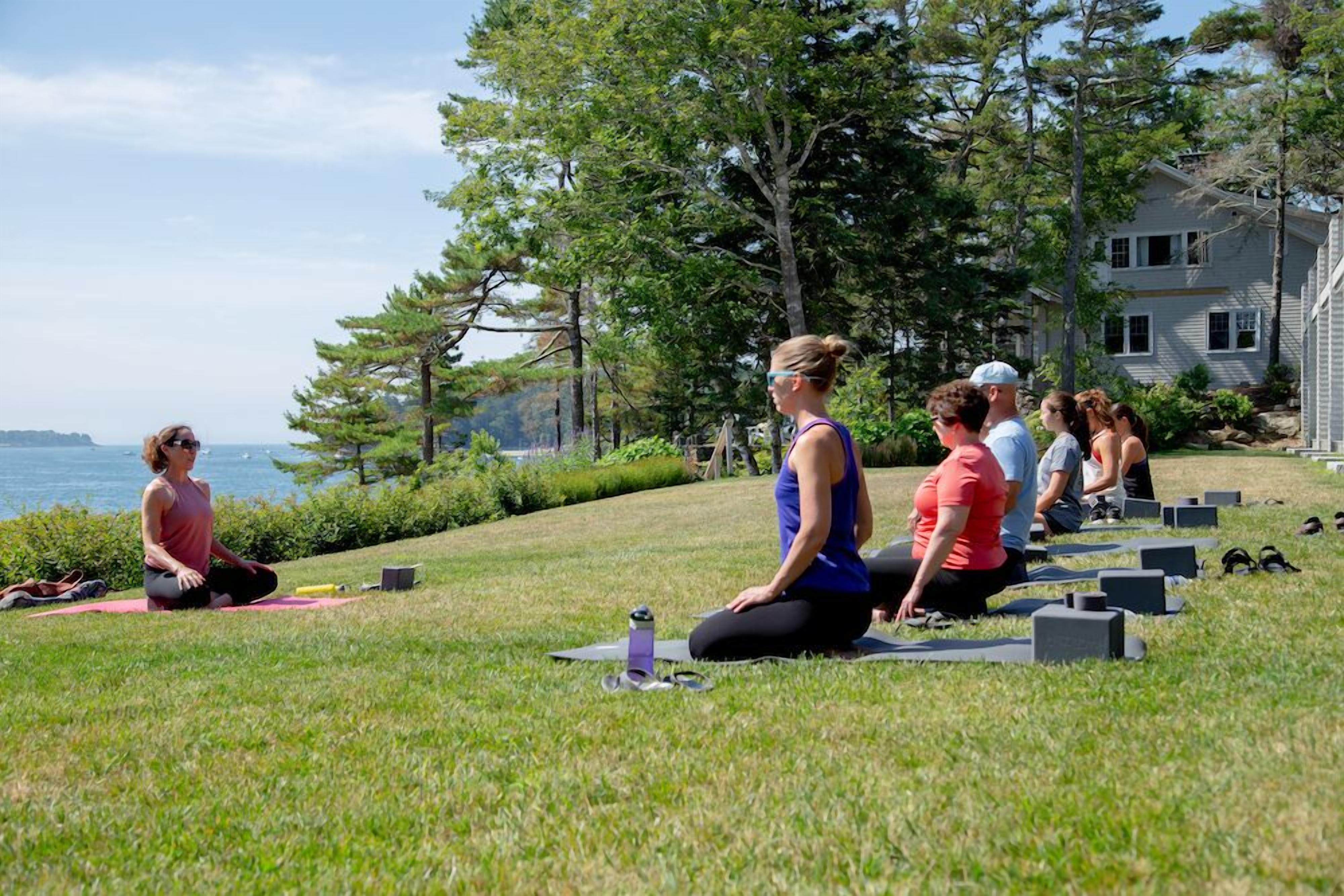 A group of people performing yoga on grass near a body of water and trees.