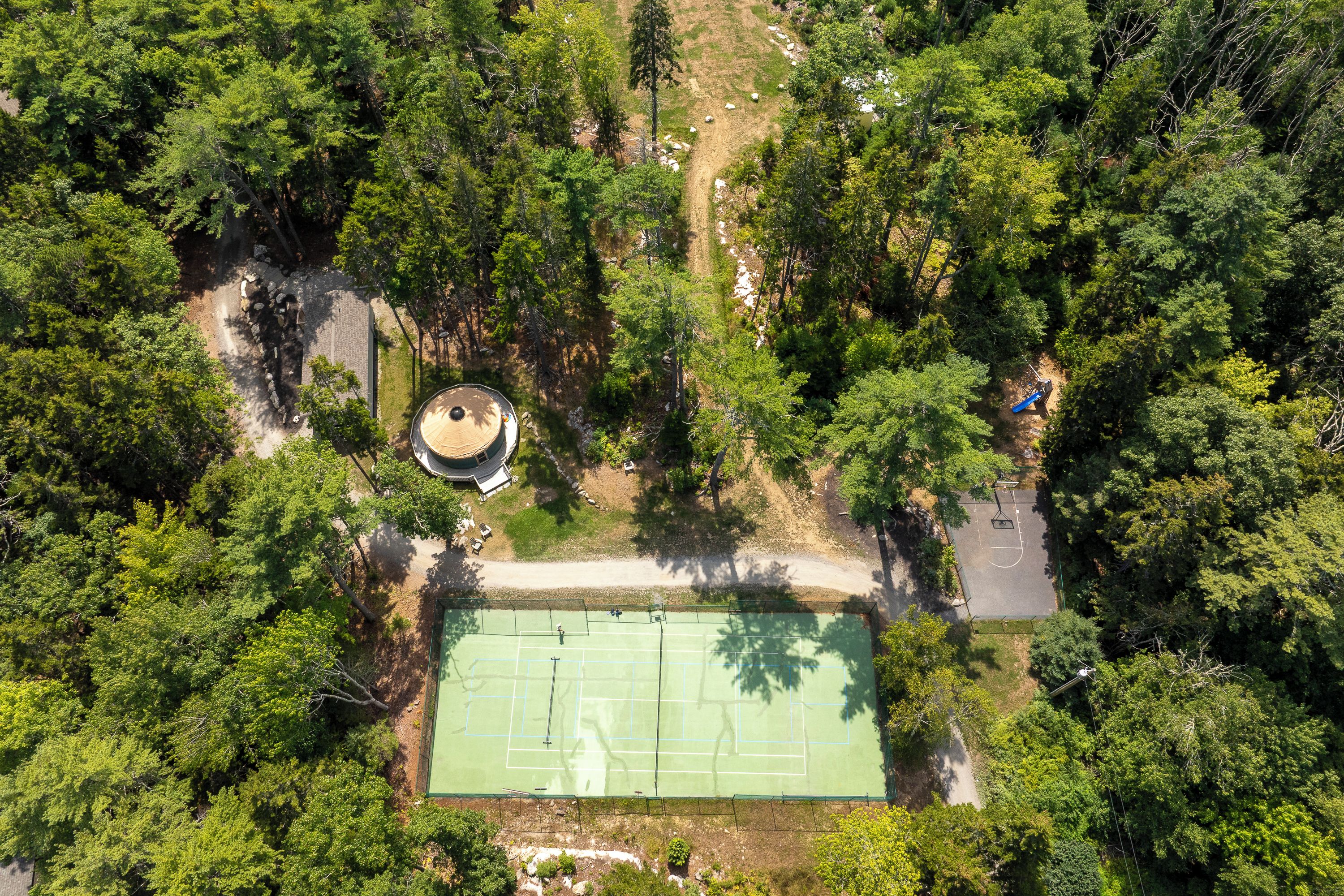 Ein Luftbild zeigt einen Tennisplatz, der von dichtem Wald umgeben ist, mit einem kleinen Gebäude und einem Feldweg in der Nähe.