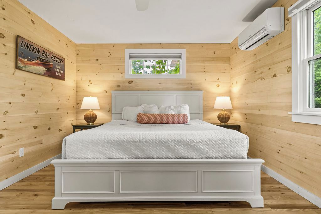 Une chambre avec un mur en bois, un lit avec une literie blanche, deux lampes de chevet, une fenêtre et un appareil de climatisation.