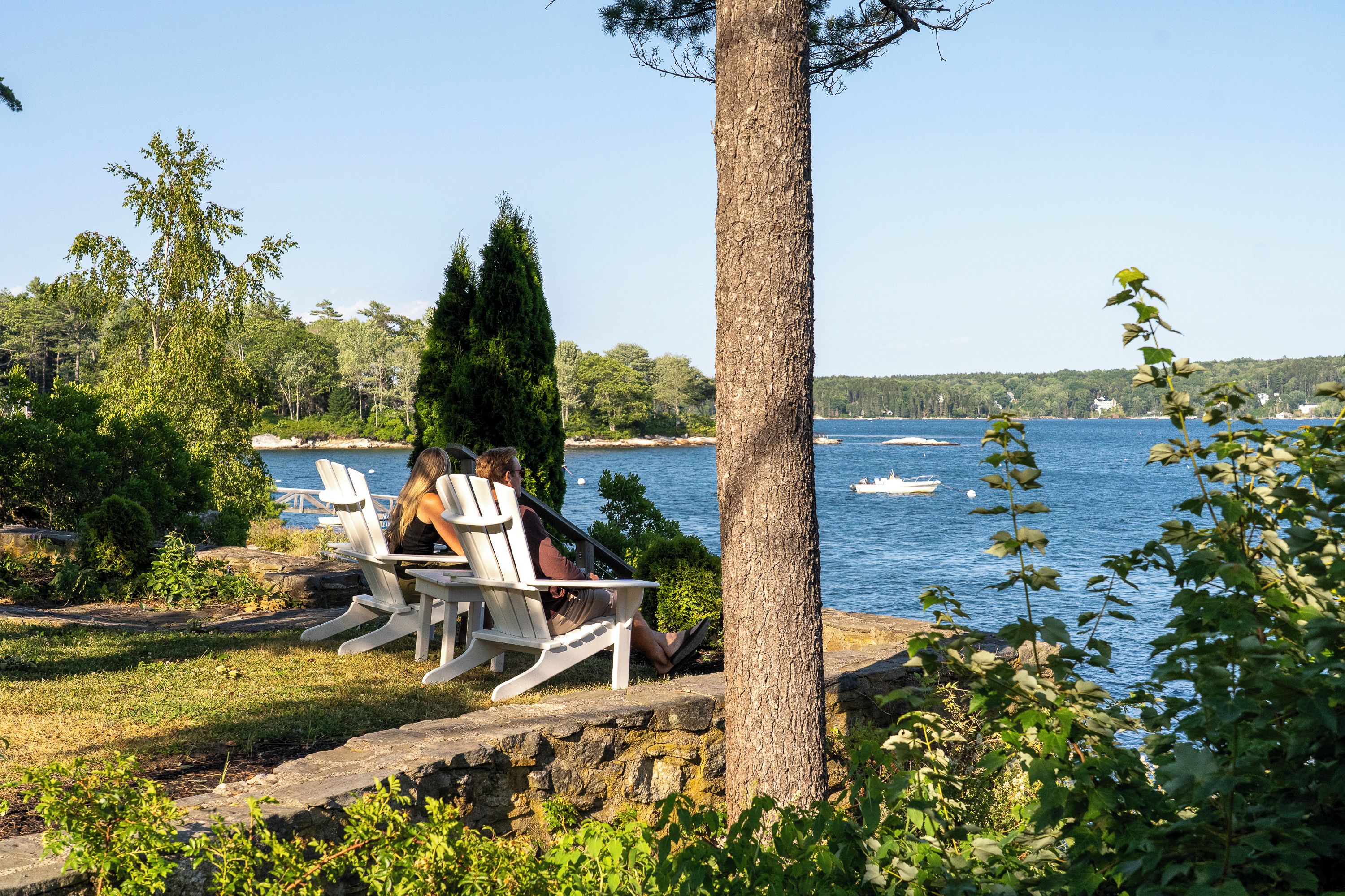 A lakeside setting with two people seated at a table, surrounded by greenery and a view of the water.