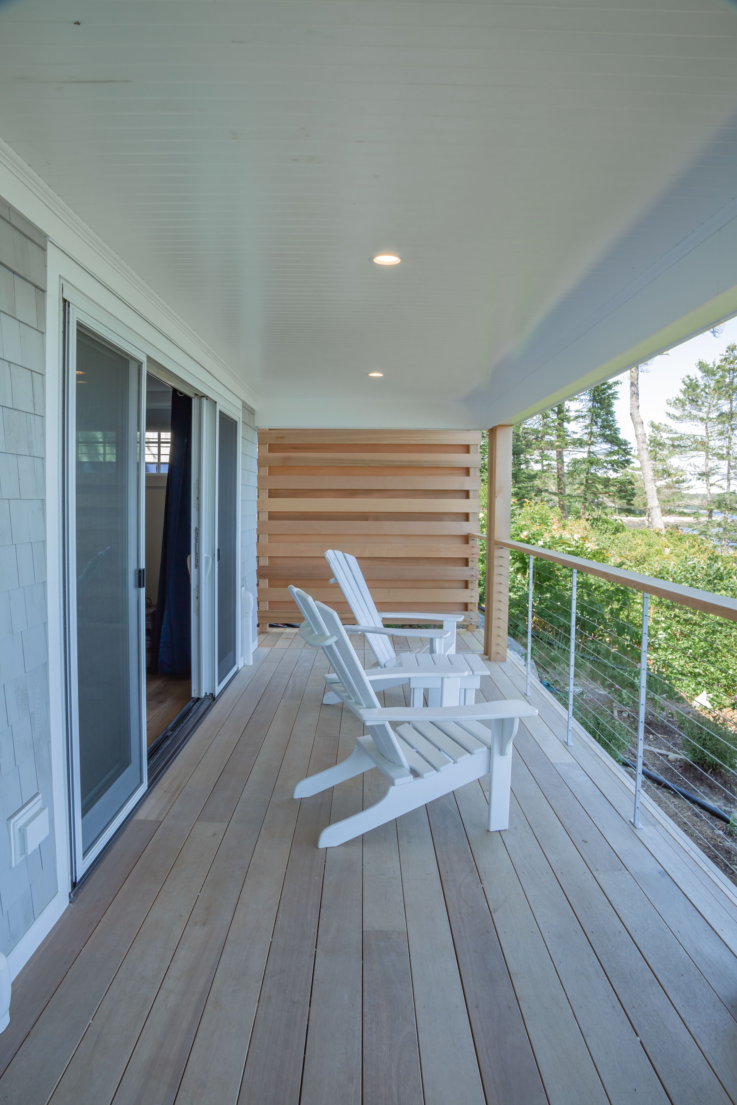 A wooden deck with white chairs, a sliding glass door, and a view of greenery.