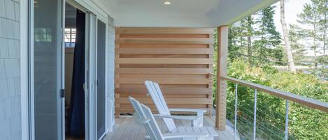 A wooden deck with white chairs, a sliding glass door, and a view of greenery.