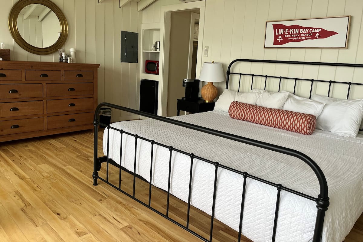Une chambre avec un lit en métal, une commode en bois et une enseigne fixée au mur indiquant « Lin-E-Kin Bay Camp ».