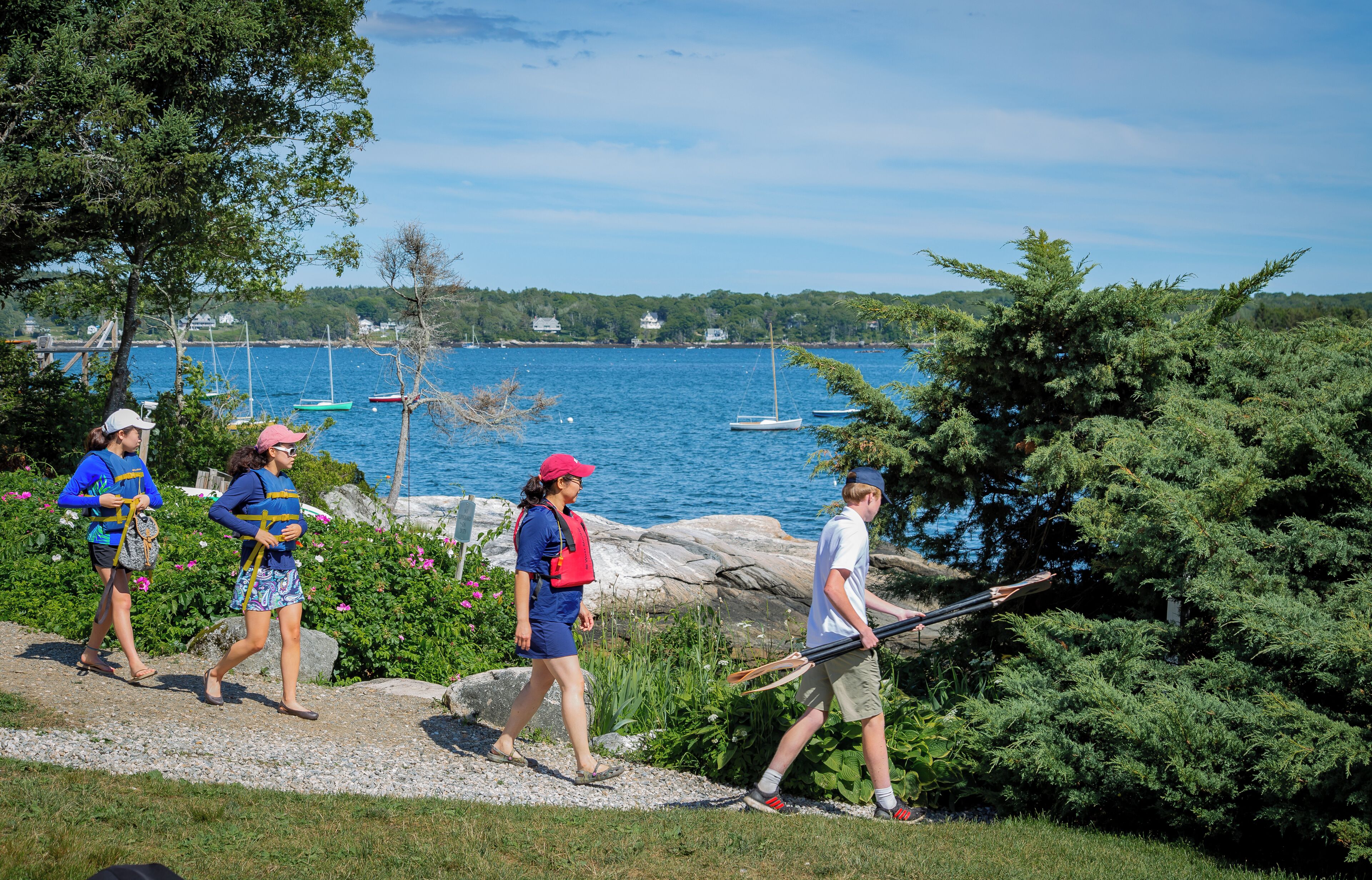 A group of people walking on a path near a body of water with boats in the background.