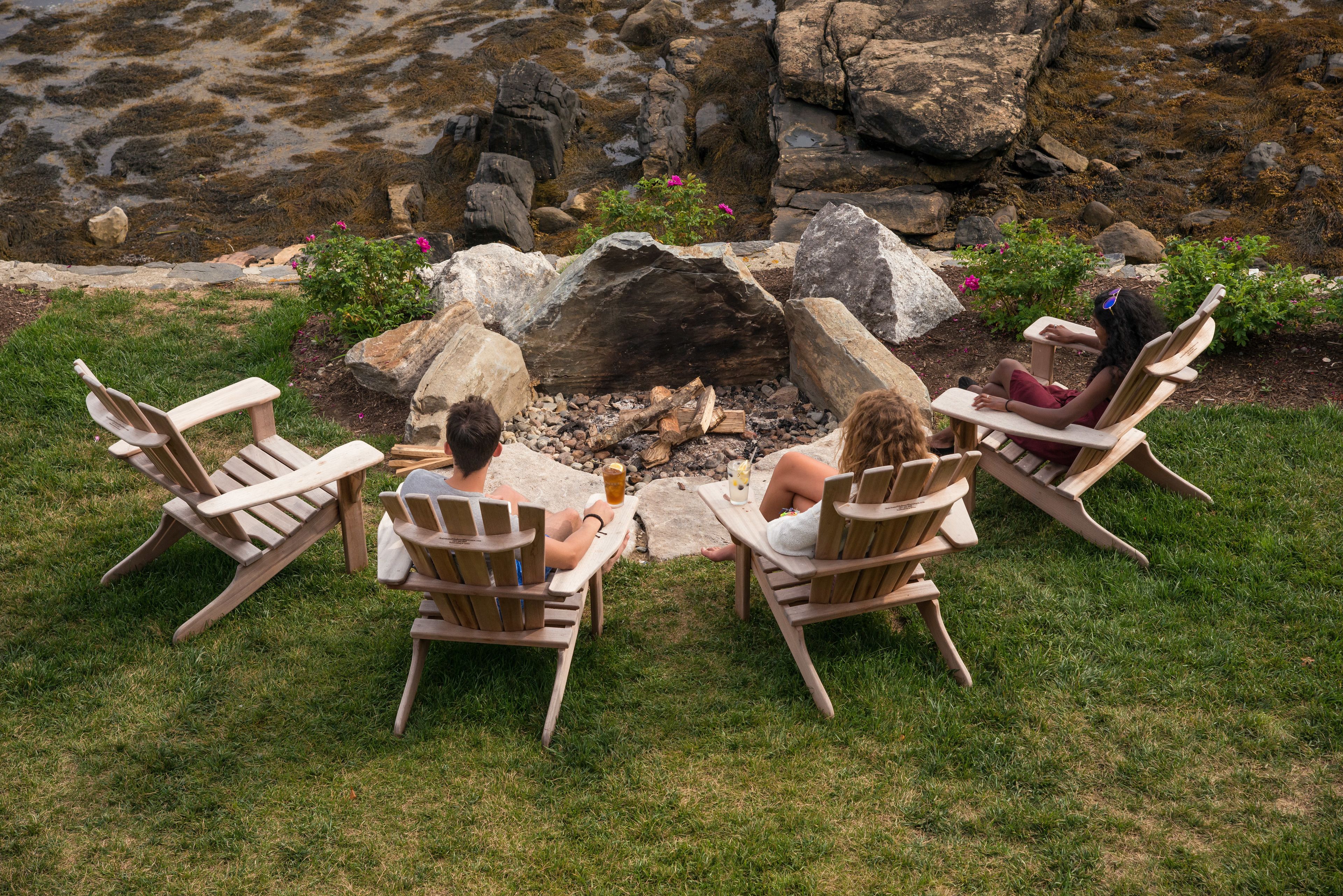 Three wooden Adirondack chairs around a stone fire pit in a grassy outdoor area with rocks and flowers.