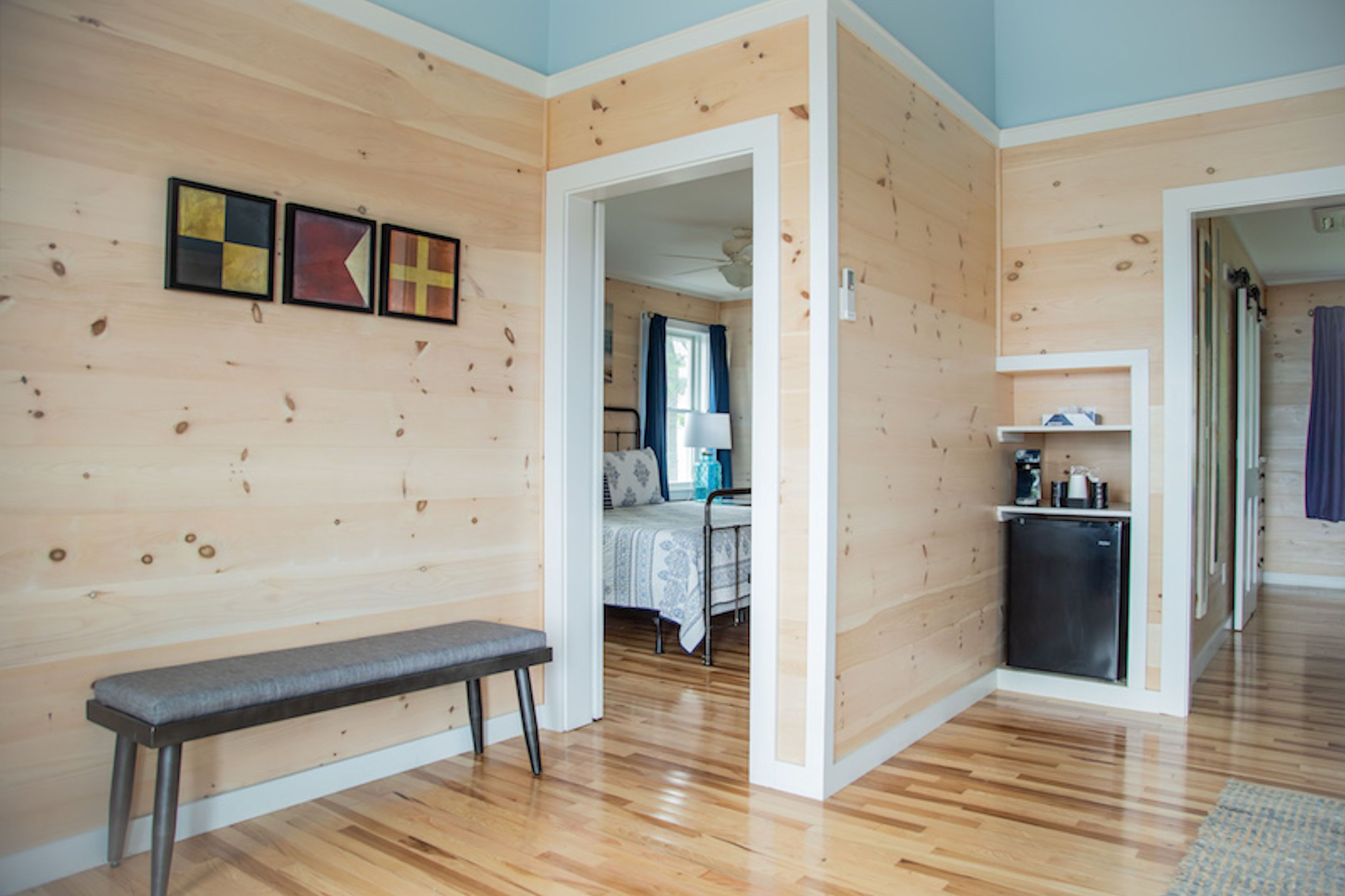 A room with wooden walls and floors, a bench, a bed with a blue and white bedspread, a small kitchenette with a fridge, and a door leading to another room.
