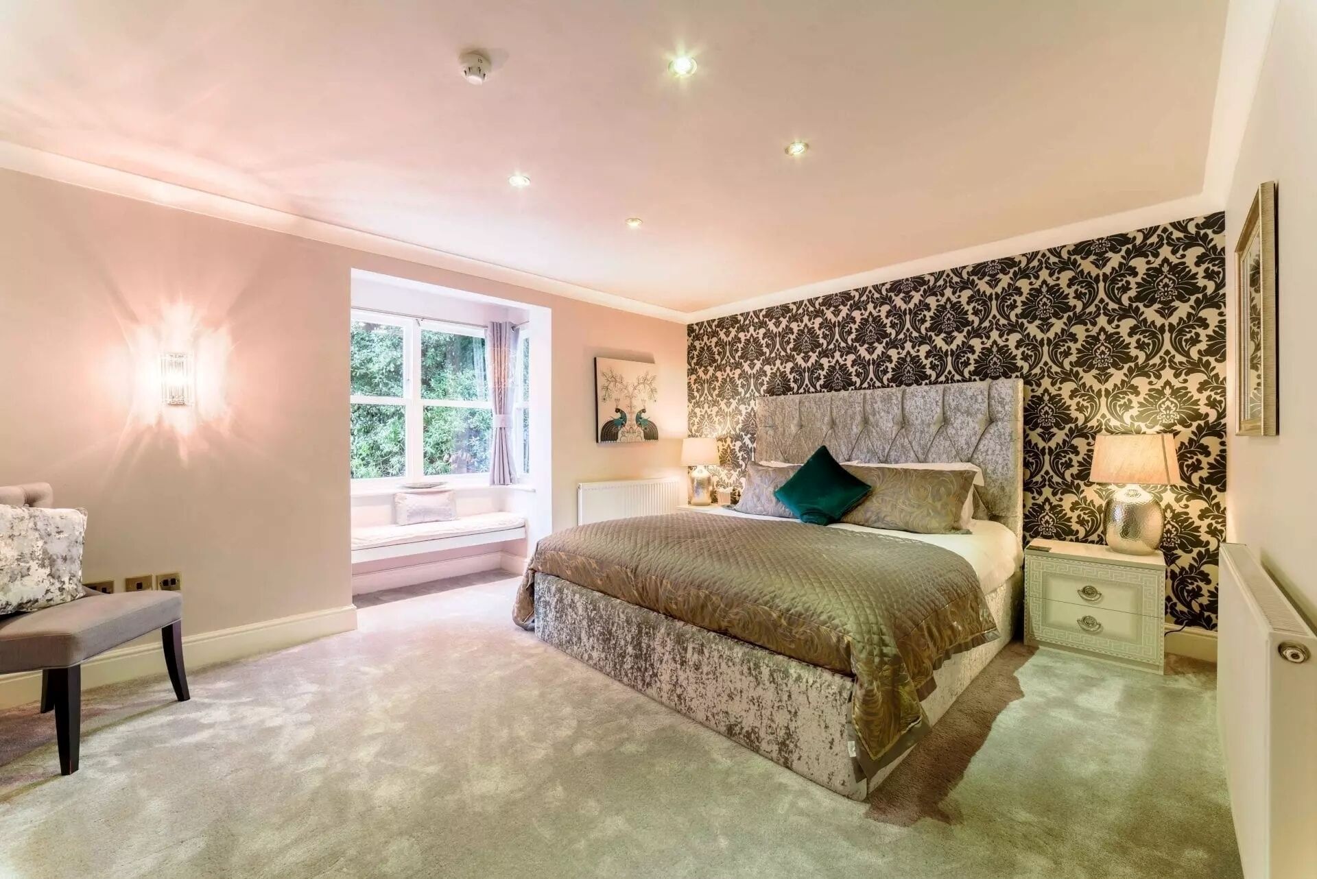A bedroom with a large bed, a bench, a window with curtains, and patterned wallpaper.