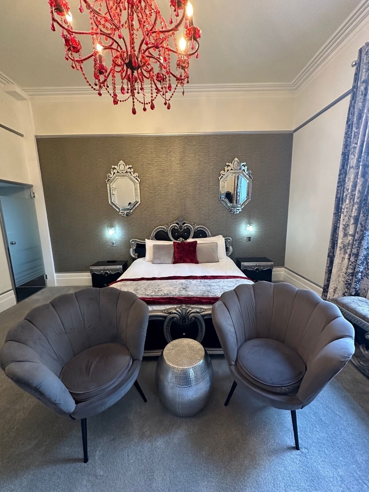 A bedroom with a bed, two armchairs, a chandelier, and two wall mirrors.